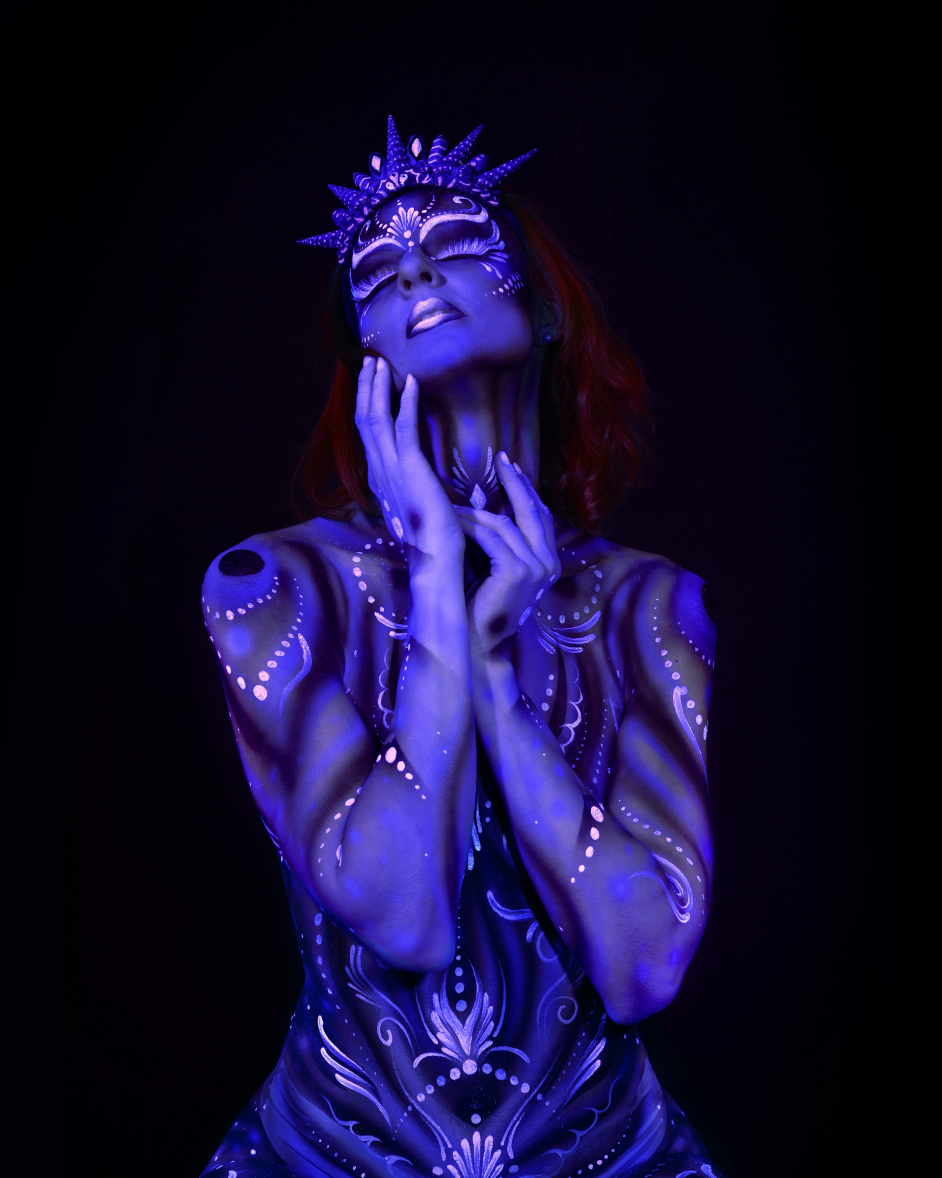 Julez stands with her head tilted back and her eyes closed. A decorative pattern in blue and purple with dotted and scroll details covers her torso, complemented by a spiked crown headpiece. Bodypaint by Joana Rohr. Photographed by Elad Itzkin Photography under blacklight in his Gesundbrunnen, Berlin studio.