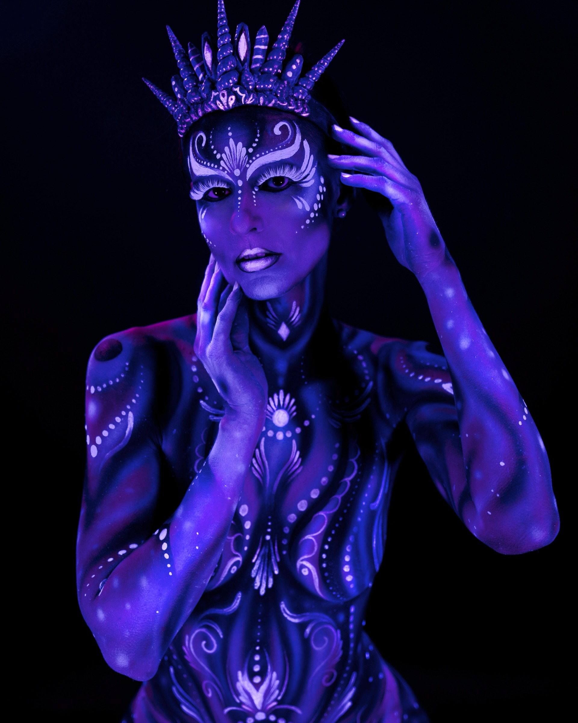 Julez stands facing the camera directly. Her body is covered in an ornamental blue and purple design with dotted and floral style linework, and she wears a spiked crown headpiece. Bodypaint by Joana Rohr. Photographed by Elad Itzkin Photography under UV light in his Gesundbrunnen, Berlin studio.