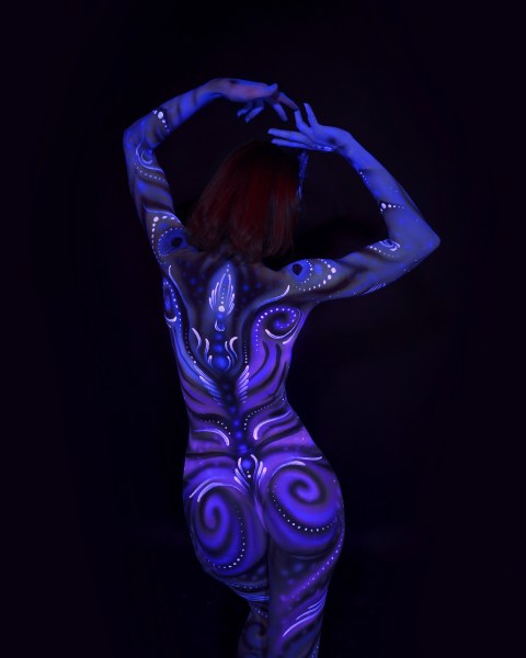 Julez is photographed with her back to the camera. A symmetrical ornamental design in blue and purple, featuring swirls and dotted linework, covers her back. Bodypaint by Joana Rohr. Photographed by Elad Itzkin Photography under UV blacklight in his Gesundbrunnen, Berlin studio.