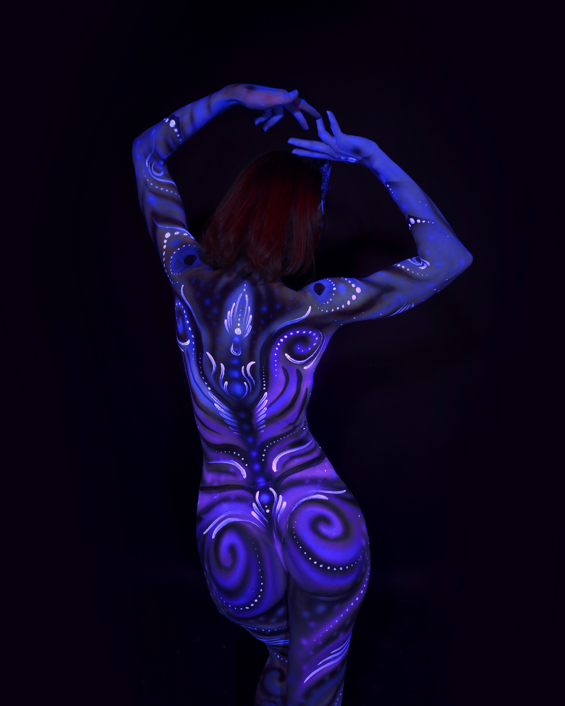 Julez is photographed with her back to the camera. A symmetrical ornamental design in blue and purple, featuring swirls and dotted linework, covers her back. Bodypaint by Joana Rohr. Photographed by Elad Itzkin Photography under UV blacklight in his Gesundbrunnen, Berlin studio.