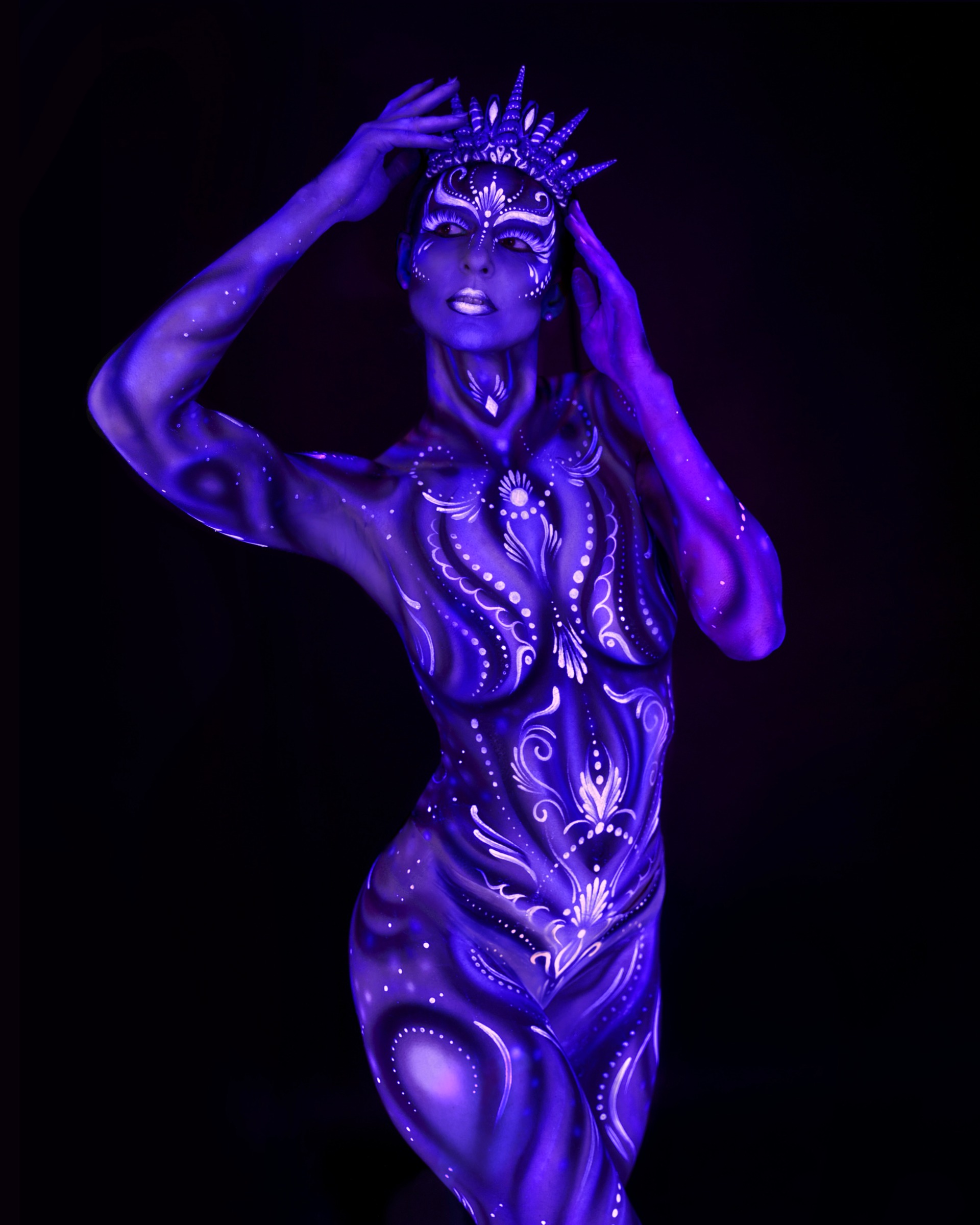 Julez stands facing the camera, wearing a spiked crown headpiece. An ornamental blue and purple pattern with dotted and floral details covers her torso. Bodypaint by Joana Rohr. Photographed by Elad Itzkin Photography under UV light in his Gesundbrunnen, Berlin studio.