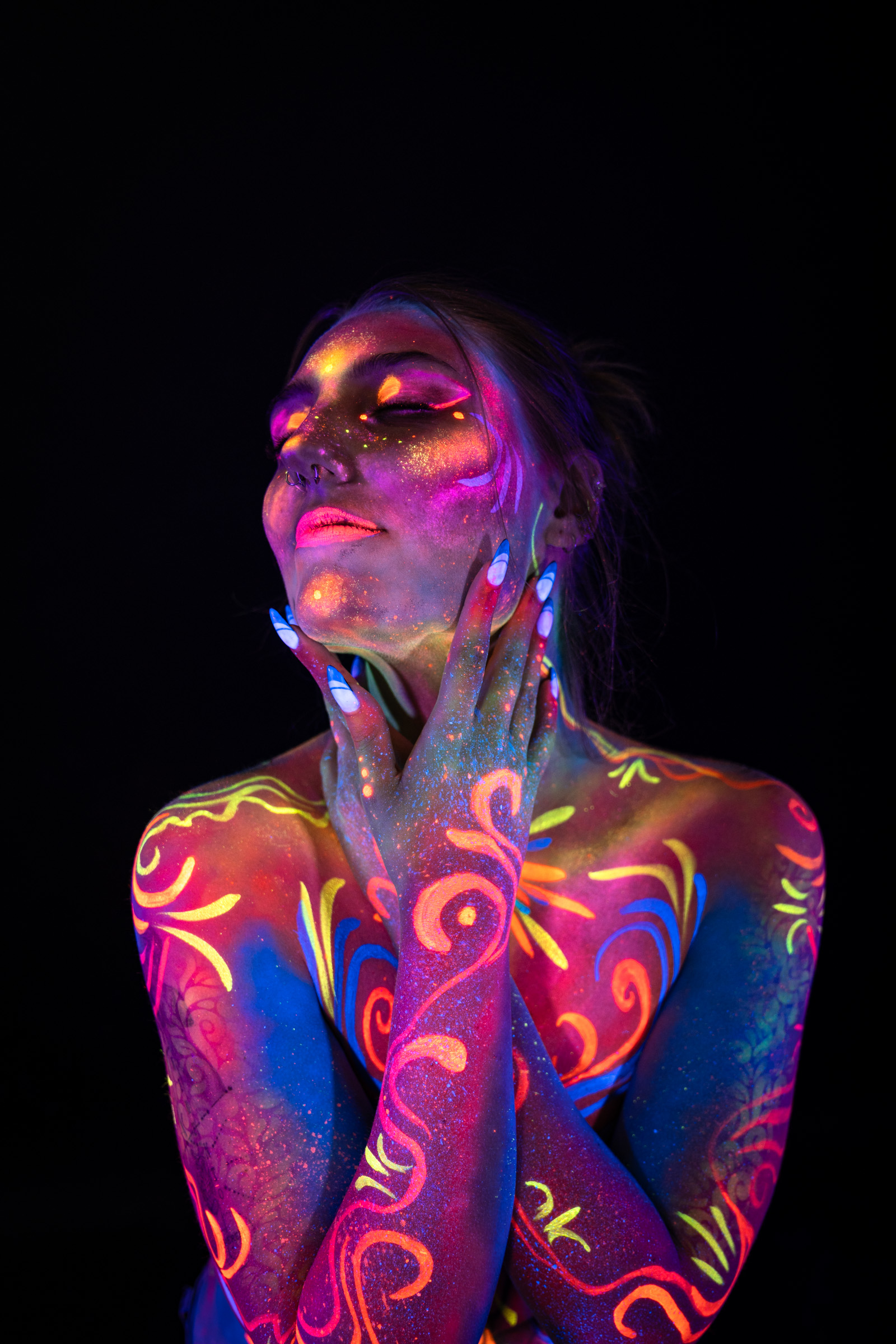 Kiki is shown from the chest up, head tilted back and turned to one side in a three-quarter view. The bodypaint is a swirling design in pink, orange, blue, and yellow tones across the face, neck, and shoulders. Bodypaint by Elad Itzkin, Sage Brewer, and Hanni. Photographed by Elad Itzkin Photography under UV light in his studio in Gesundbrunnen, Berlin.