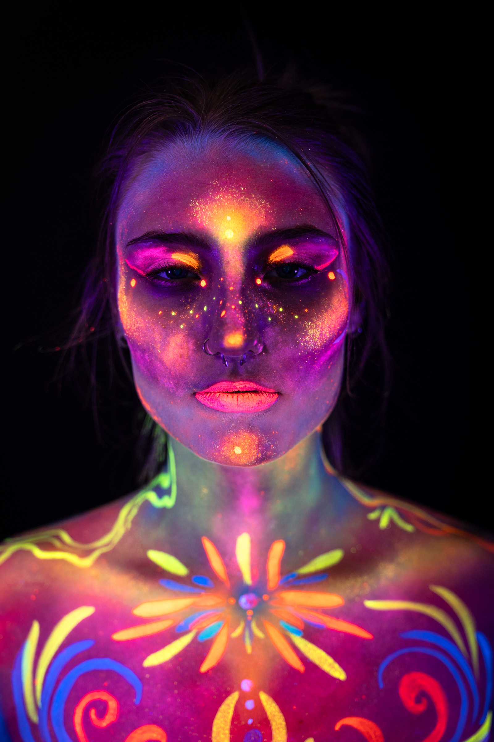 Kiki is framed from the chest up, facing the camera directly. Her face and shoulders are covered in a colorful swirl pattern combining pink, orange, blue, and yellow, with a starburst motif centered on the chest. Bodypaint by Elad Itzkin, Sage Brewer, and Hanni. Photographed by Elad Itzkin Photography under blacklight in his studio in Gesundbrunnen, Berlin.