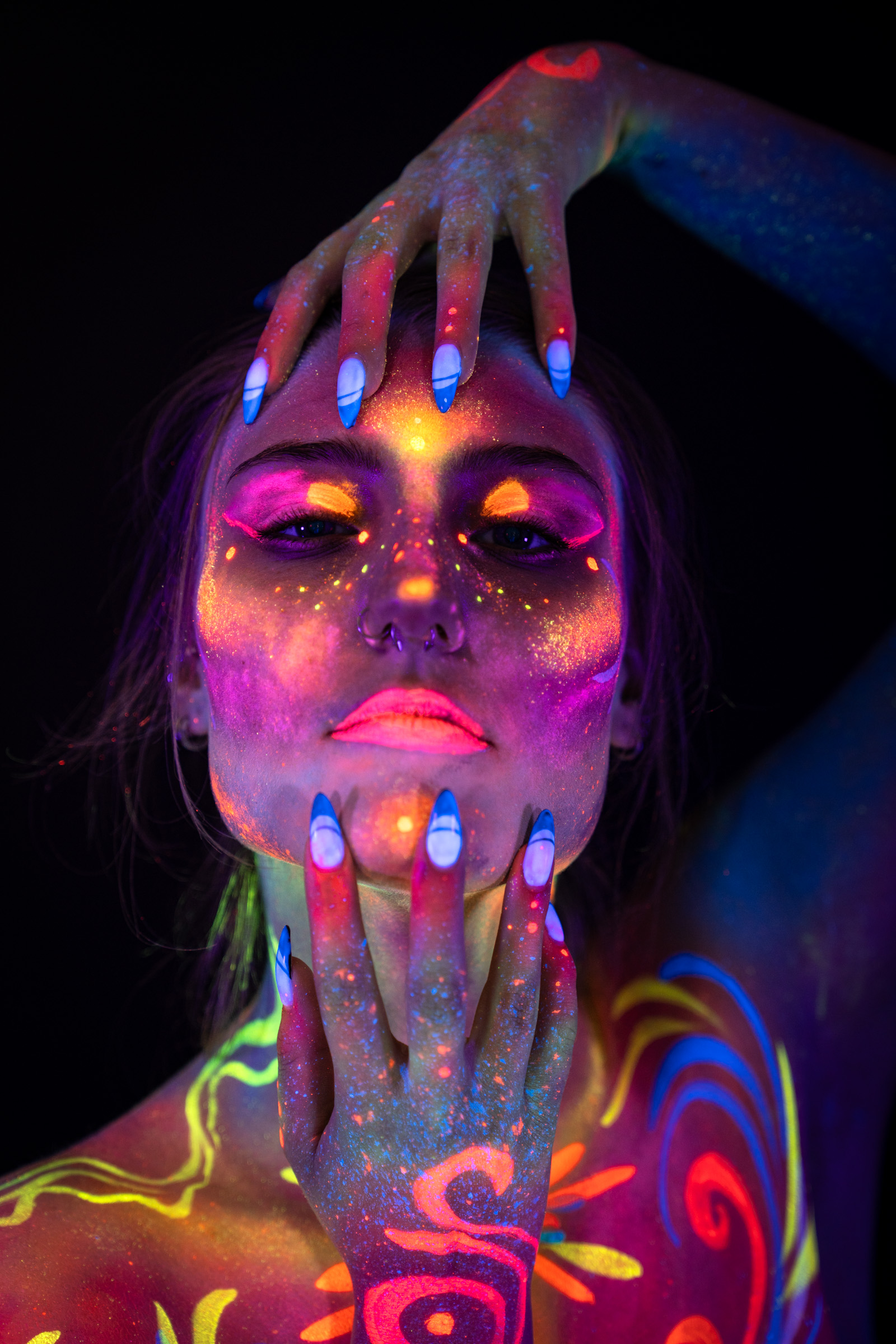 Kiki is shown from the chest up, facing forward with a direct gaze. A swirling bodypaint pattern in pink, orange, blue, and yellow covers her face, neck, and shoulders. Bodypaint by Elad Itzkin, Sage Brewer, and Hanni. Photographed by Elad Itzkin Photography under UV light in his studio in Gesundbrunnen, Berlin.