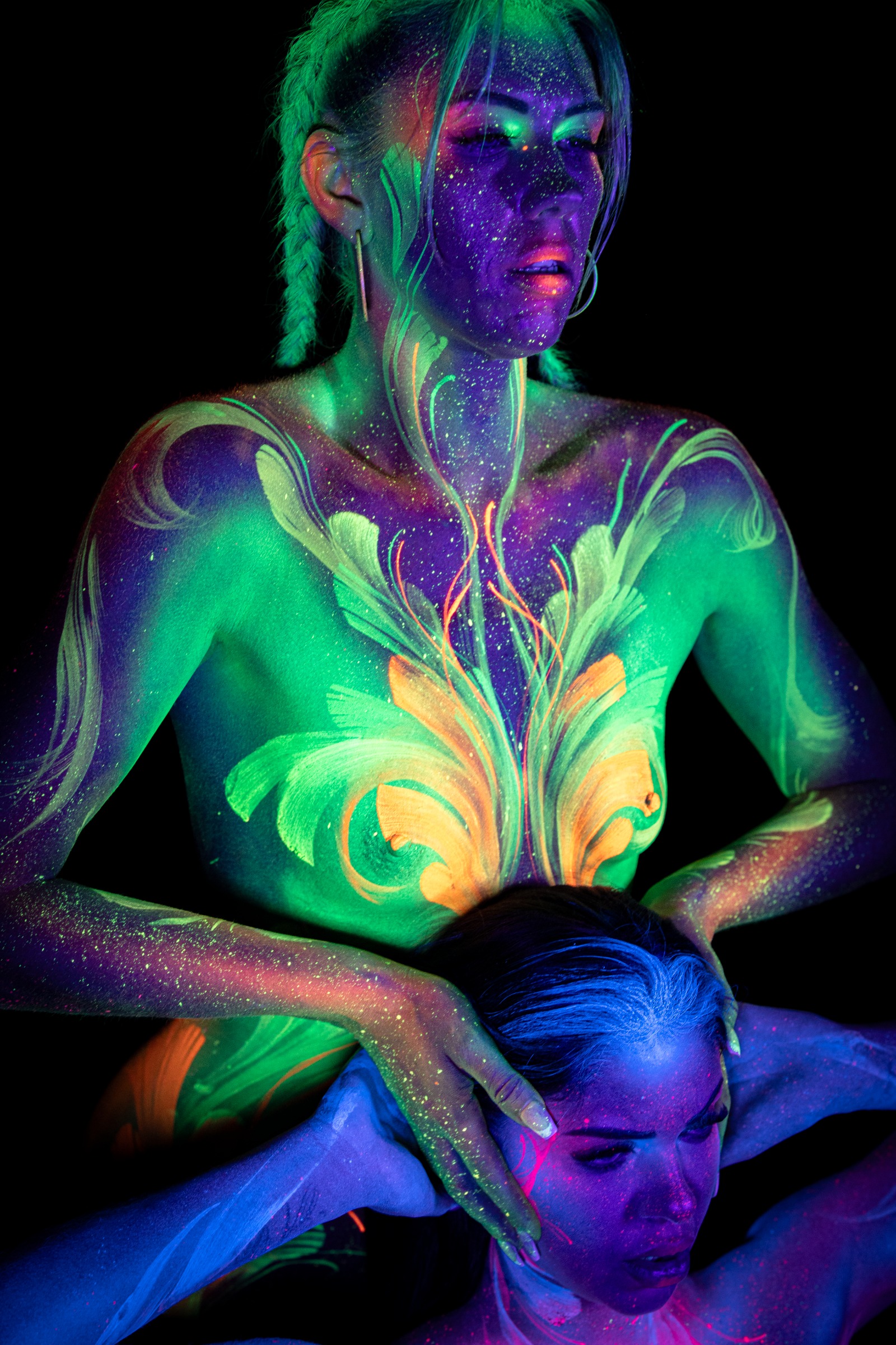 LadySnke and Miss_Layla_X pose together, LadySnke in front with her head tilted back and Miss_Layla_X positioned behind her. LadySnke wears a pink and white floral design, Miss_Layla_X a green and orange floral linework pattern. Bodypaint by Claudia Kraemer. Photographed by Elad Itzkin Photography under UV blacklight in Gesundbrunnen, Berlin.