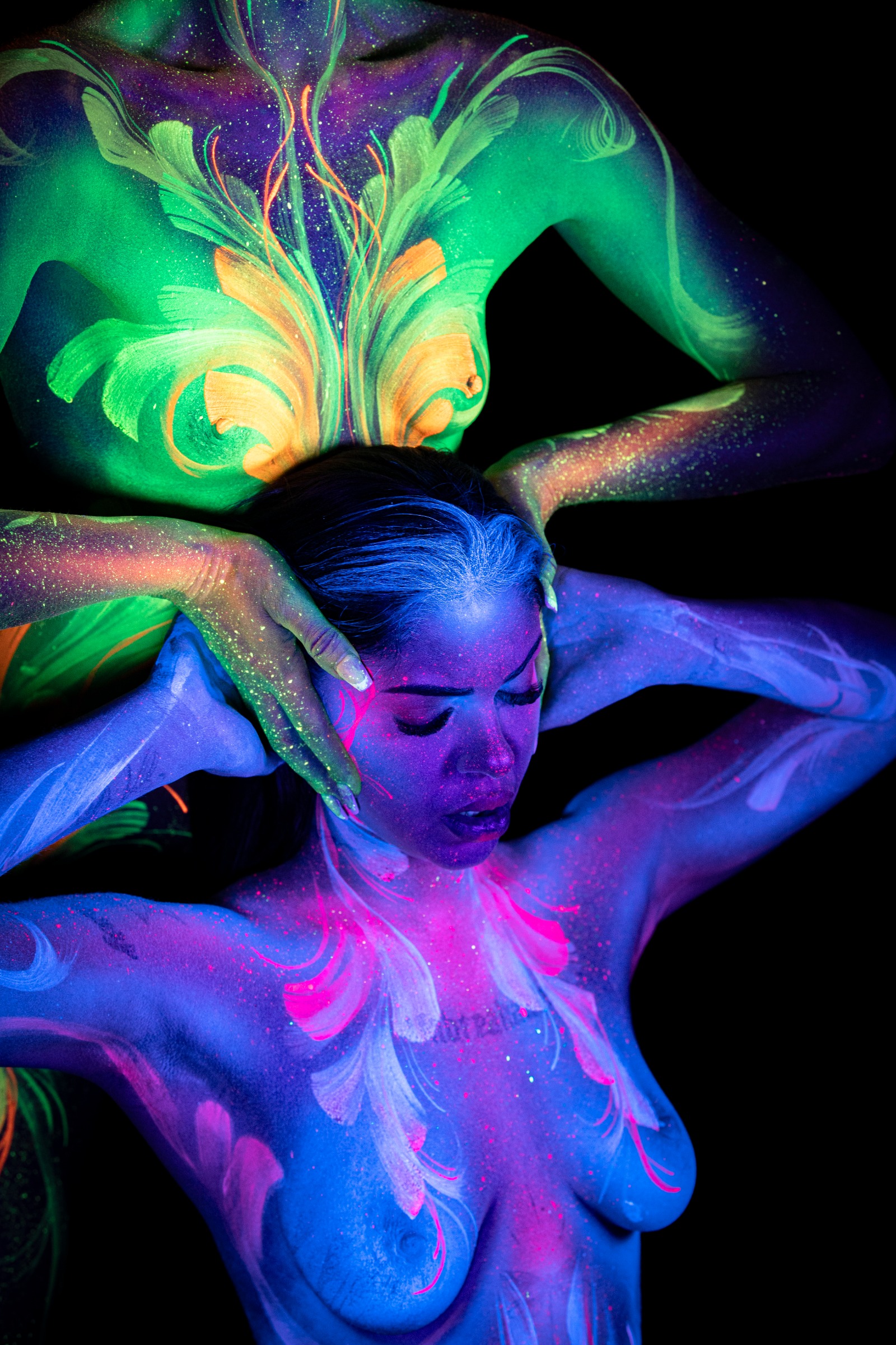 Miss_Layla_X stands behind LadySnke, who tilts her head back in the foreground. Miss_Layla_X's torso shows a green and orange floral linework design, while LadySnke's chest carries a pink and white pattern. Bodypaint by Claudia Kraemer. Photographed by Elad Itzkin Photography under UV blacklight in his Gesundbrunnen, Berlin studio.