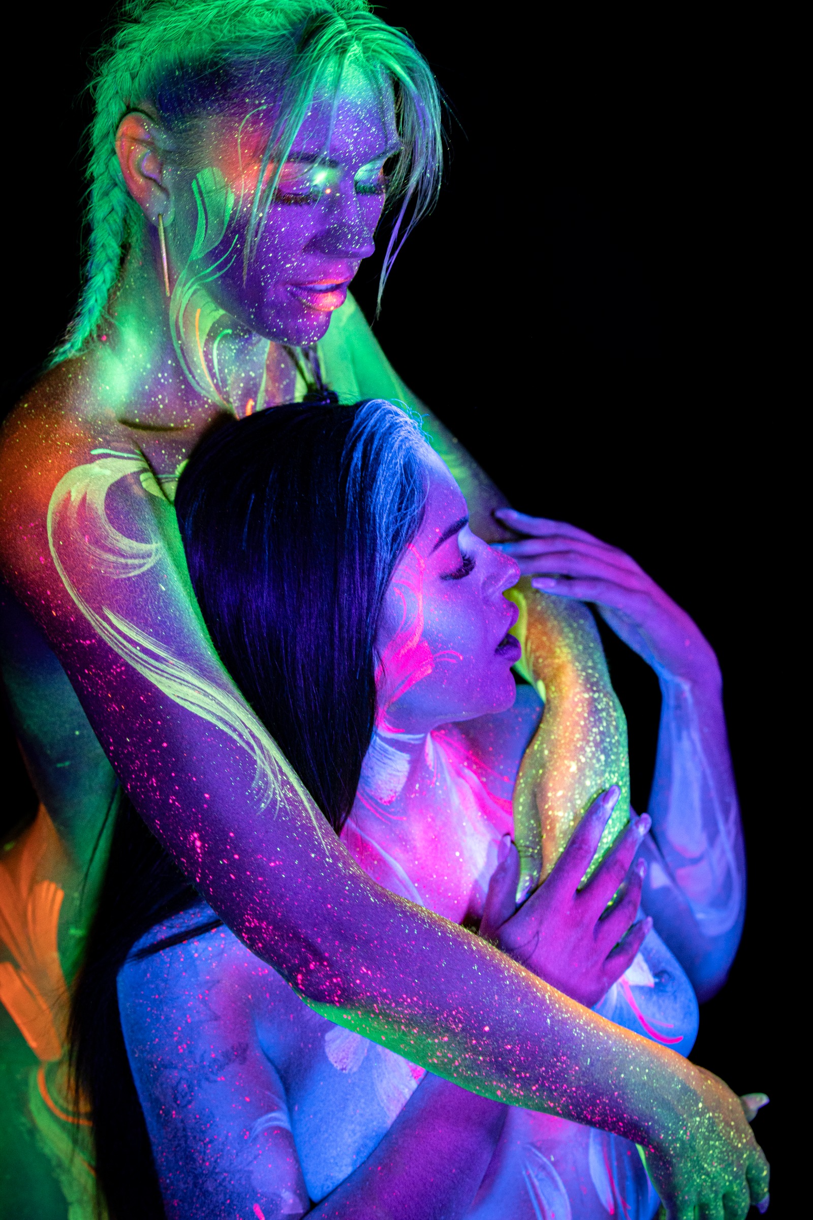 Miss_Layla_X and LadySnke pose close together facing each other, Miss_Layla_X's face turned down toward LadySnke in front of her. Green and orange floral linework and pink and white linework mark their torsos respectively. Bodypaint by Claudia Kraemer. Photographed by Elad Itzkin Photography under UV blacklight in his Gesundbrunnen, Berlin studio.