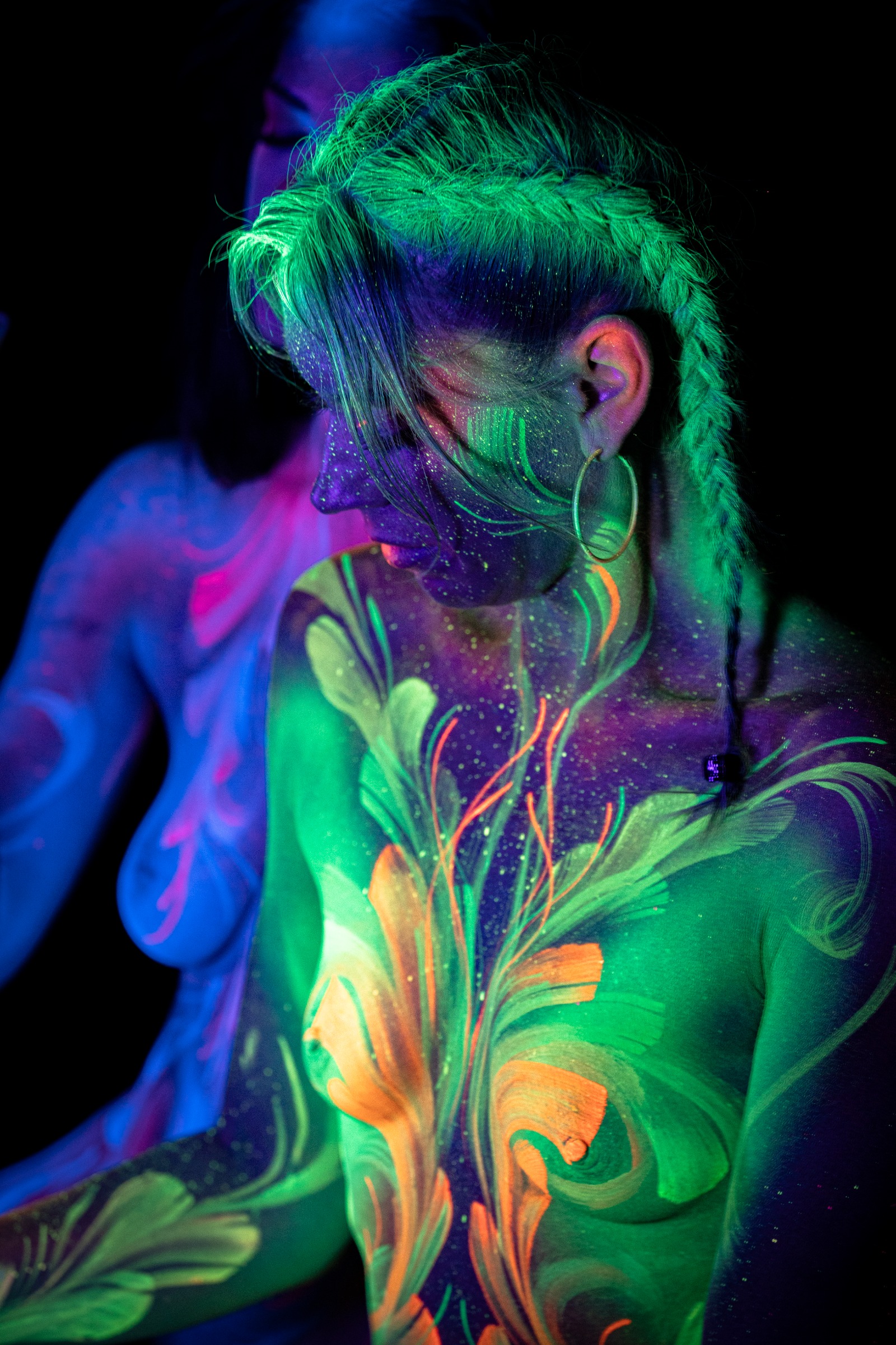 Miss_Layla_X and LadySnke are seated close together facing the same direction, their heads near one another. Green and orange floral linework and pink and white linework decorate their torsos. Bodypaint by Claudia Kraemer. Photographed by Elad Itzkin Photography under UV blacklight in his Gesundbrunnen, Berlin studio.