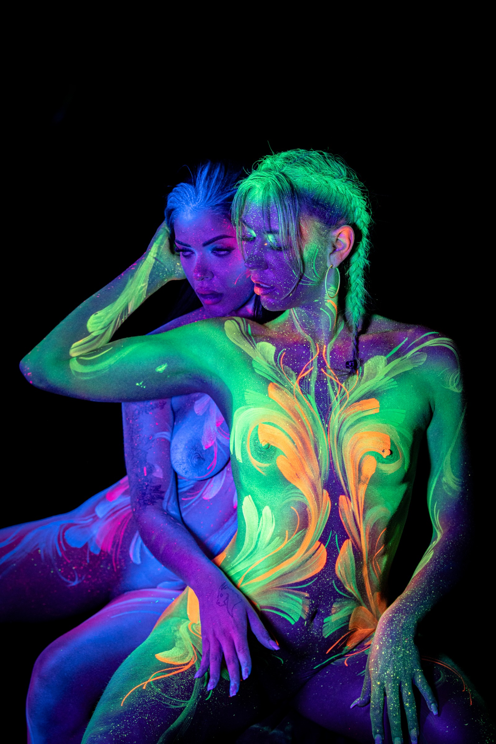 LadySnke and Miss_Layla_X are shown in a close upper-body view, seated together with their heads close and faces angled toward each other. Their bodypaint includes green and orange floral linework and a pink and white pattern. Bodypaint by Claudia Kraemer. Photographed by Elad Itzkin Photography under UV blacklight in Gesundbrunnen, Berlin.