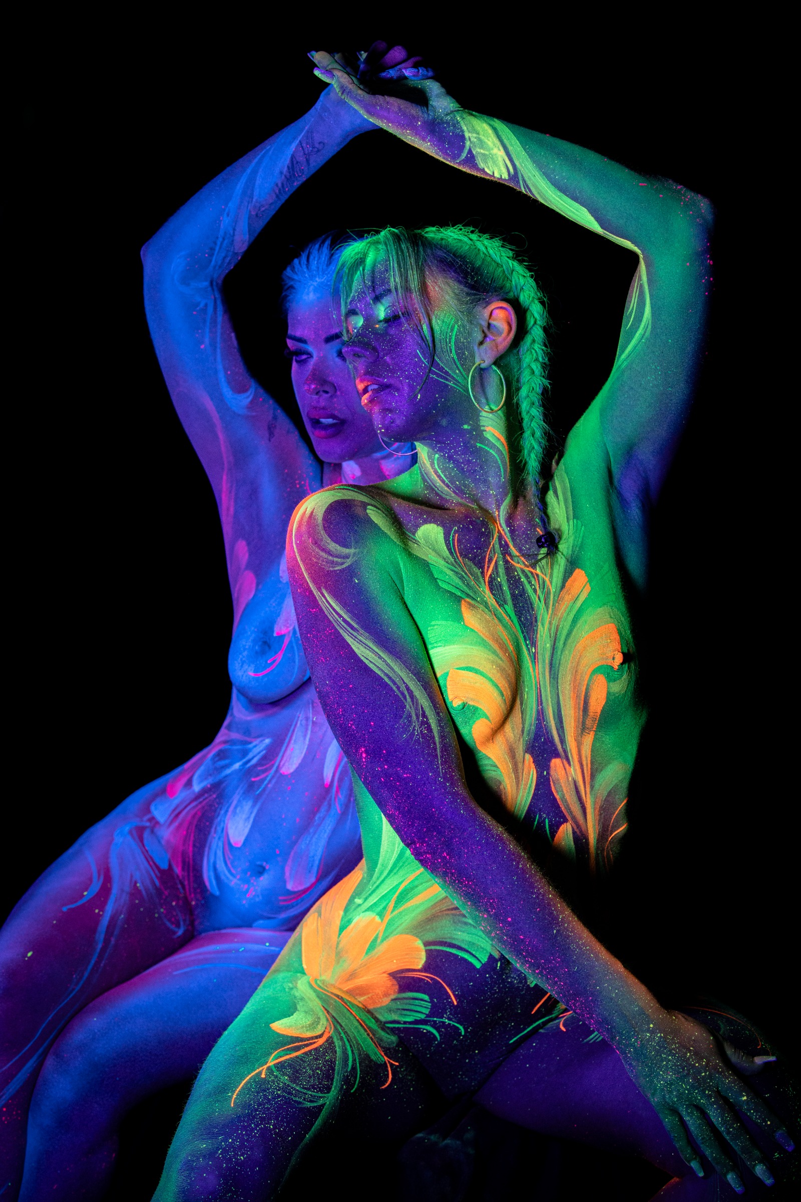 LadySnke and Miss_Layla_X sit close together in a three-quarter view, heads tilted toward one another. The bodypaint features a blue and purple swirl pattern on one model and a green, yellow and orange floral motif on the other. Bodypaint by Claudia Kraemer. Photographed by Elad Itzkin Photography under UV/blacklight in his studio in Gesundbrunnen, Berlin.