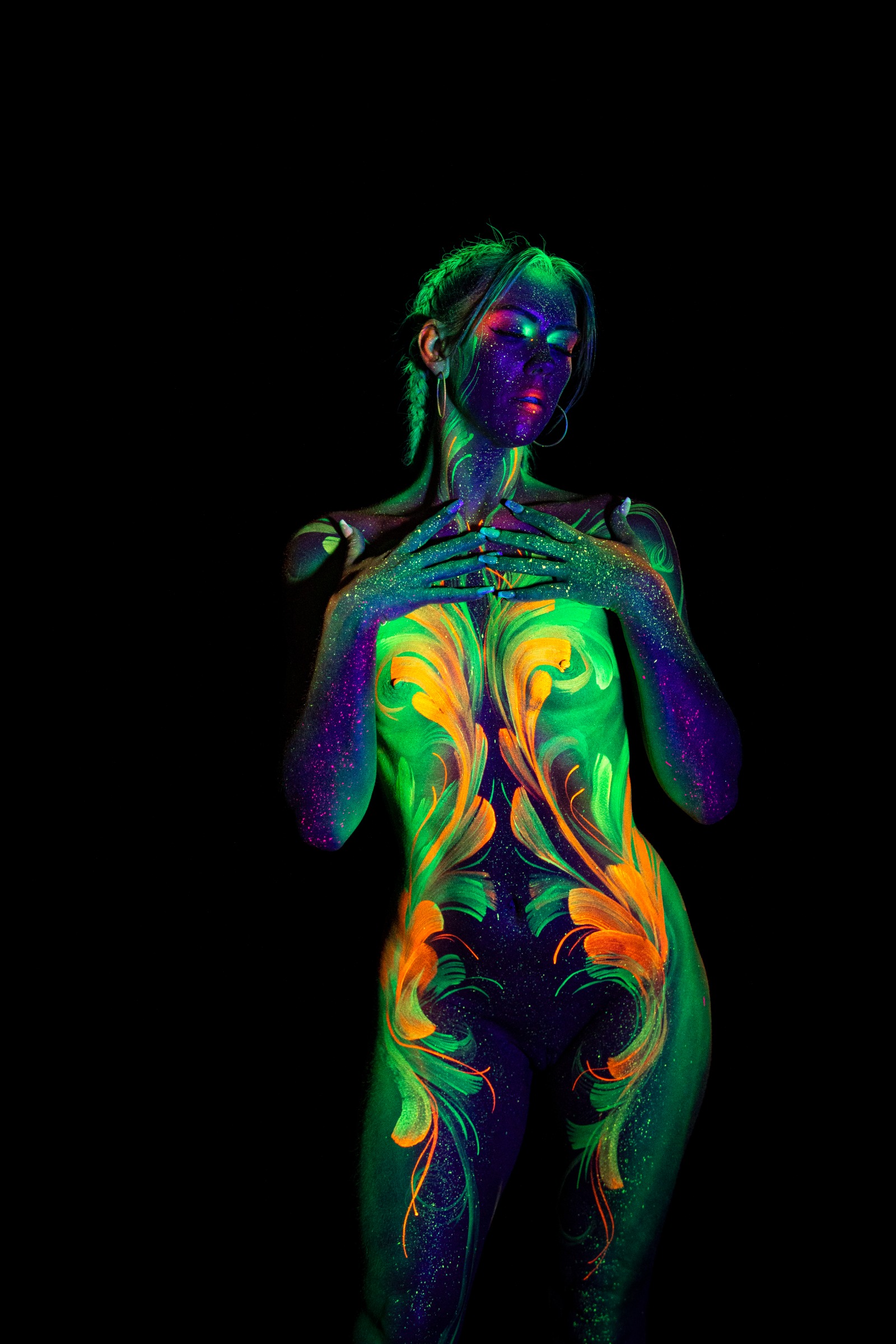 Miss_Layla_X stands facing forward toward the camera. The torso is painted with a symmetrical green and orange floral linework design, with the face rendered in deep purple. Bodypaint by Claudia Kraemer. Photographed by Elad Itzkin Photography under UV/blacklight in his studio in Gesundbrunnen, Berlin.