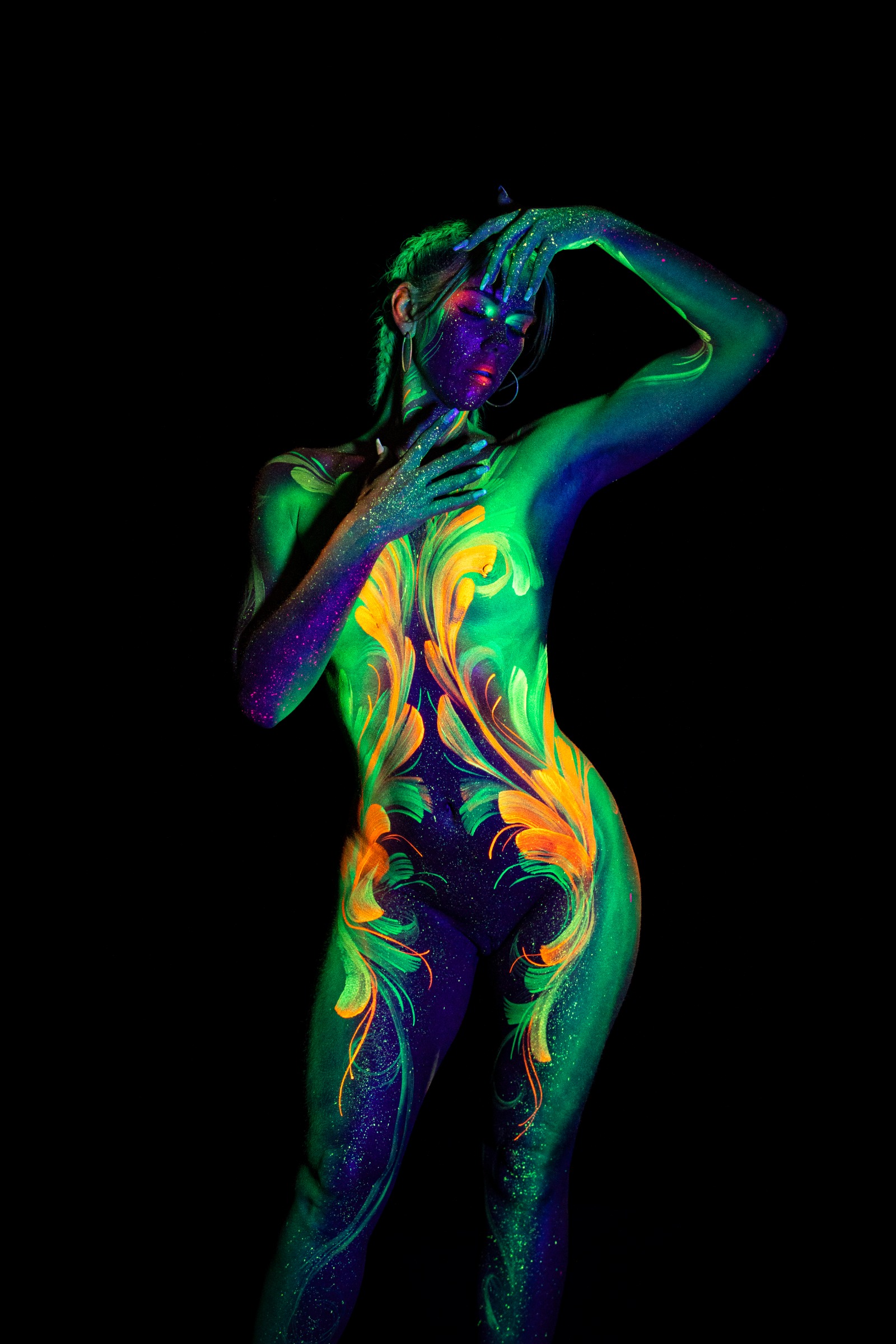 Miss_Layla_X stands in a frontal pose facing the camera. A mirrored floral linework pattern in green and orange spans the torso and continues down the legs. Bodypaint by Claudia Kraemer. Photographed by Elad Itzkin Photography under UV/blacklight in his studio in Gesundbrunnen, Berlin.