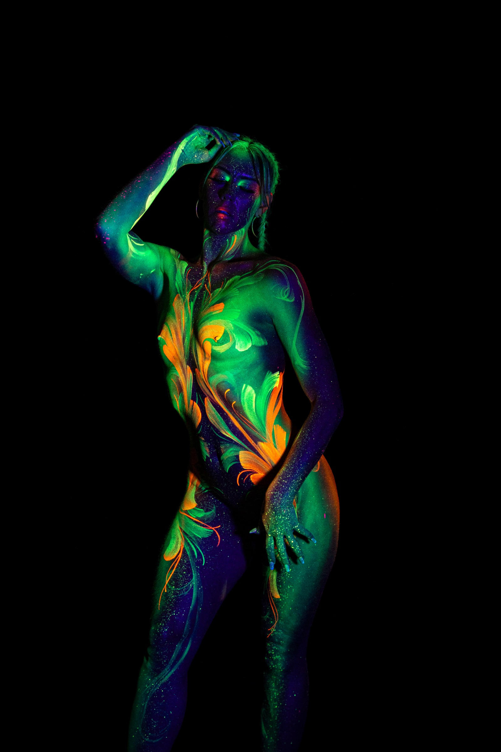 Miss_Layla_X stands facing the camera at a slight angle. Green and orange floral linework covers the chest and torso in a mirrored, symmetrical pattern. Bodypaint by Claudia Kraemer. Photographed by Elad Itzkin Photography under UV/blacklight in his studio in Gesundbrunnen, Berlin.
