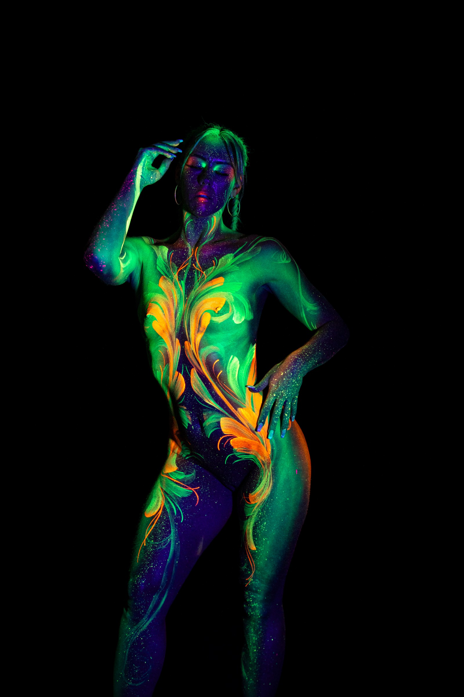 Miss_Layla_X stands with her body turned slightly to one side, facing the camera. The torso is decorated with a green and orange floral linework design on a dark purple base. Bodypaint by Claudia Kraemer. Photographed by Elad Itzkin Photography under UV/blacklight in his studio in Gesundbrunnen, Berlin.