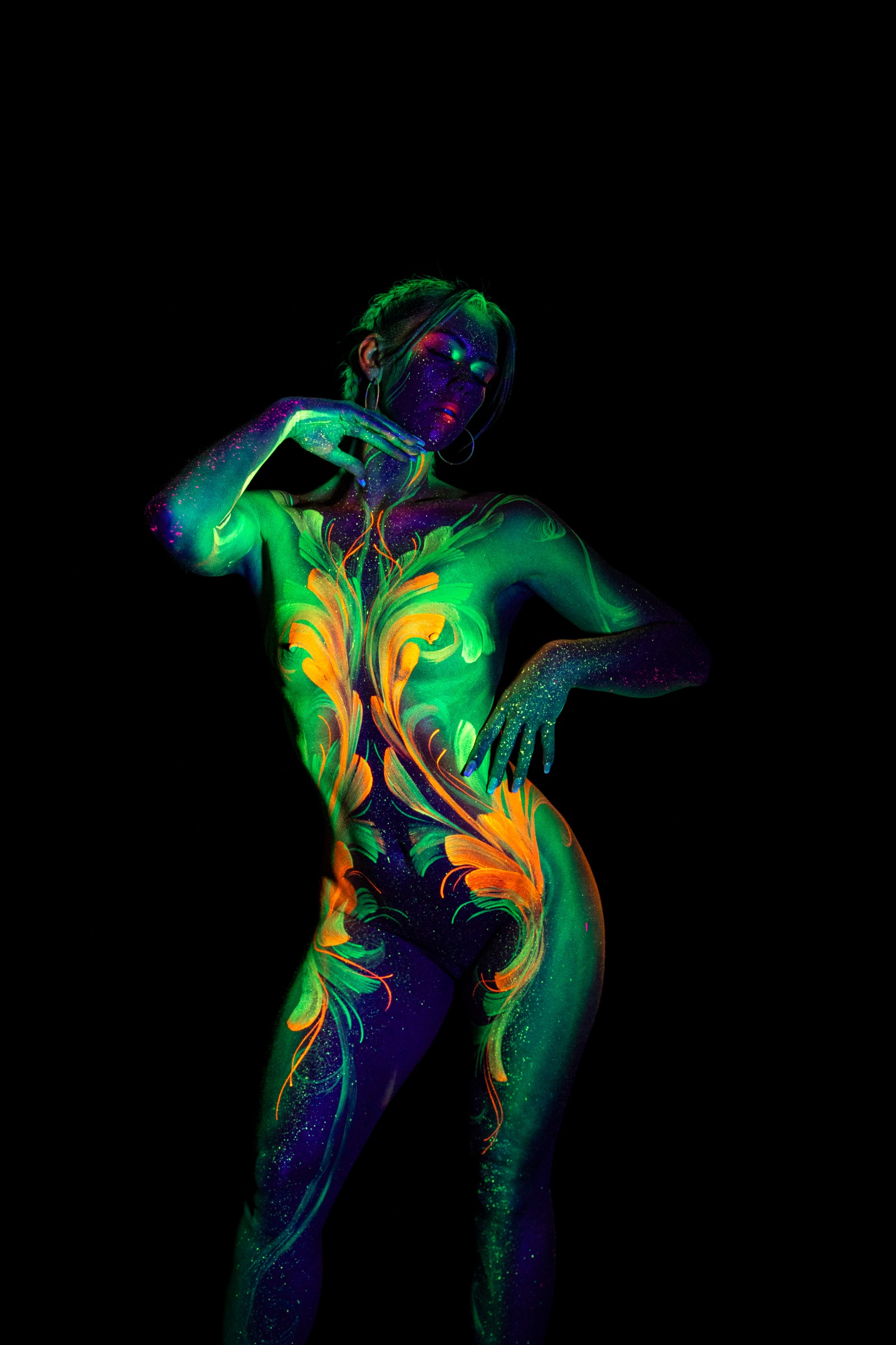 Miss_Layla_X stands facing the camera in a three-quarter view. A symmetrical green, orange and blue floral linework pattern covers the torso against a purple background tone. Bodypaint by Claudia Kraemer. Photographed by Elad Itzkin Photography under UV/blacklight in his studio in Gesundbrunnen, Berlin.