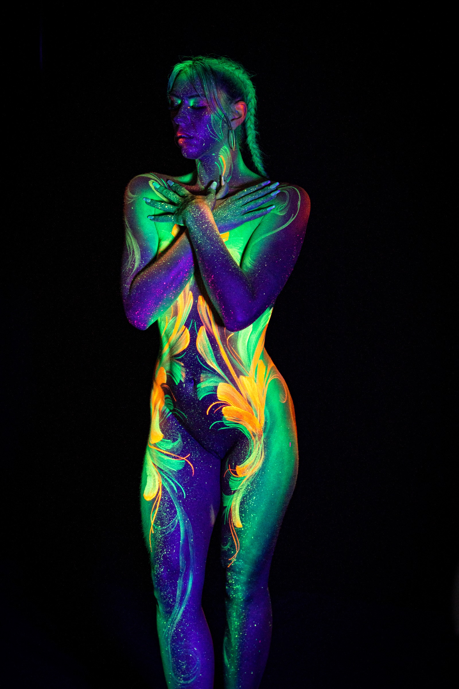 Miss_Layla_X stands upright, facing the camera directly. A green and orange botanical linework pattern spans the torso in a symmetrical, mirrored layout. Bodypaint by Claudia Kraemer. Photographed by Elad Itzkin Photography under UV/blacklight in his studio in Gesundbrunnen, Berlin.