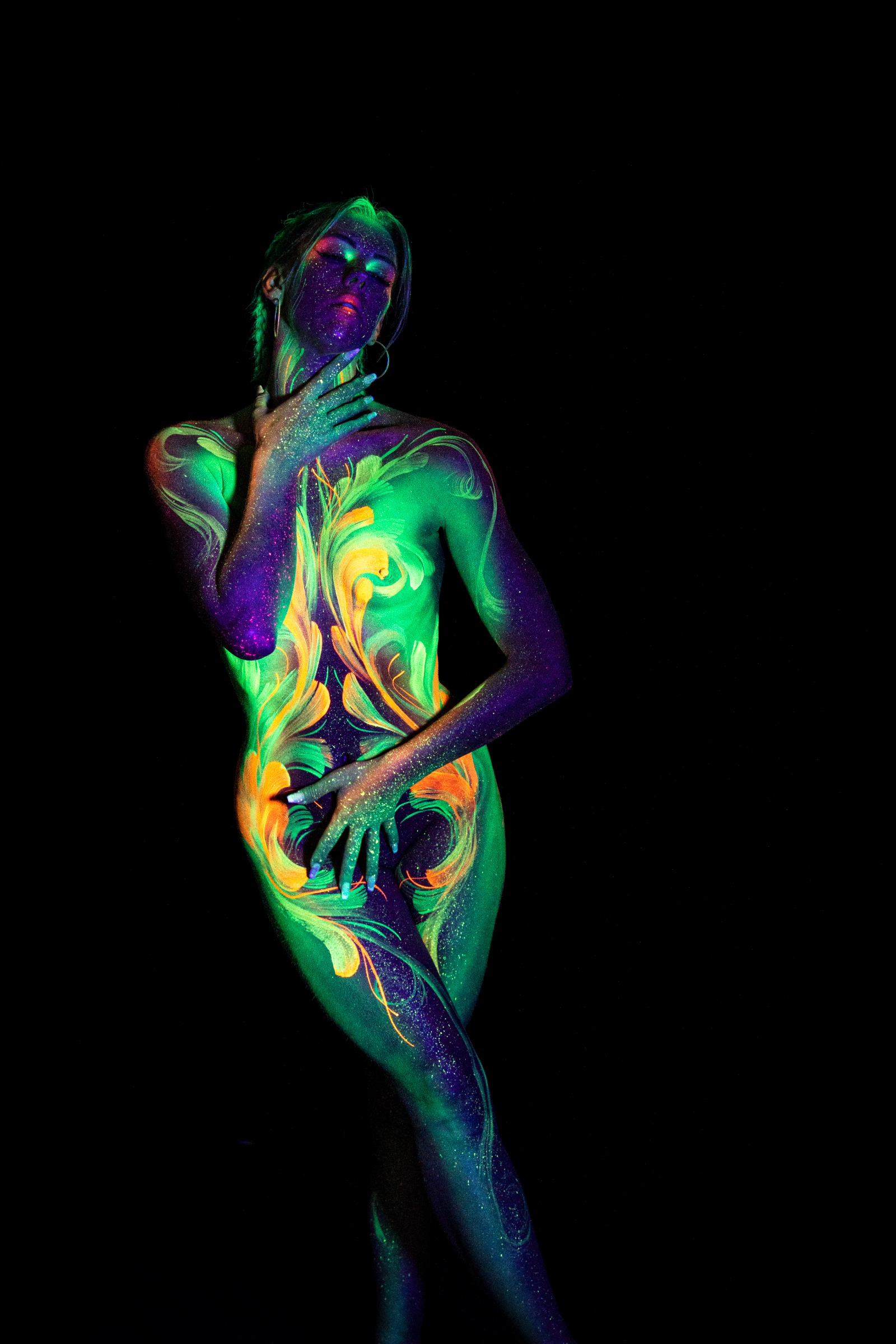 Miss_Layla_X stands with her weight on one leg in a three-quarter pose. The torso and upper leg feature a mirrored green and orange floral linework design. Bodypaint by Claudia Kraemer. Photographed by Elad Itzkin Photography under UV/blacklight in his studio in Gesundbrunnen, Berlin.