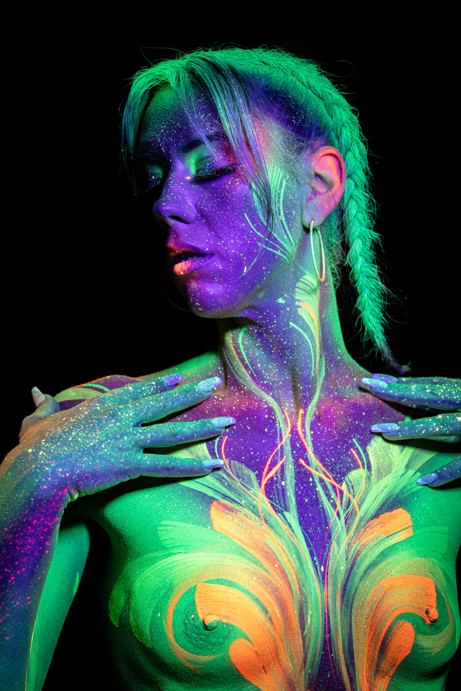 Miss_Layla_X is shown from the chest up, standing with her head tilted to one side and her eyes closed. Green and orange floral linework covers the chest in a symmetrical pattern beneath a purple painted face. Bodypaint by Claudia Kraemer. Photographed by Elad Itzkin Photography under UV/blacklight in his studio in Gesundbrunnen, Berlin.