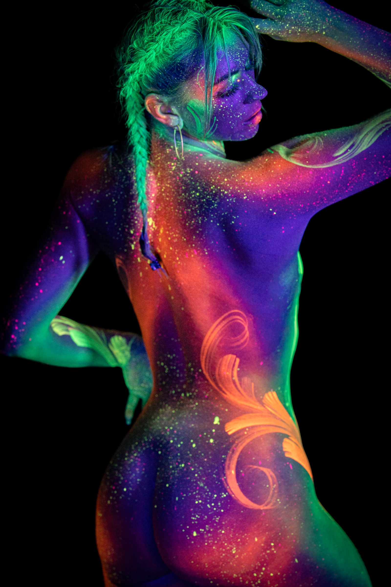 Miss_Layla_X stands with her back to the camera in a three-quarter view, an orange swirl design painted on her lower back against purple and green UV body paint. Bodypaint by Claudia Kraemer. Photographed by Elad Itzkin Photography under UV blacklight in his studio in Gesundbrunnen, Berlin.