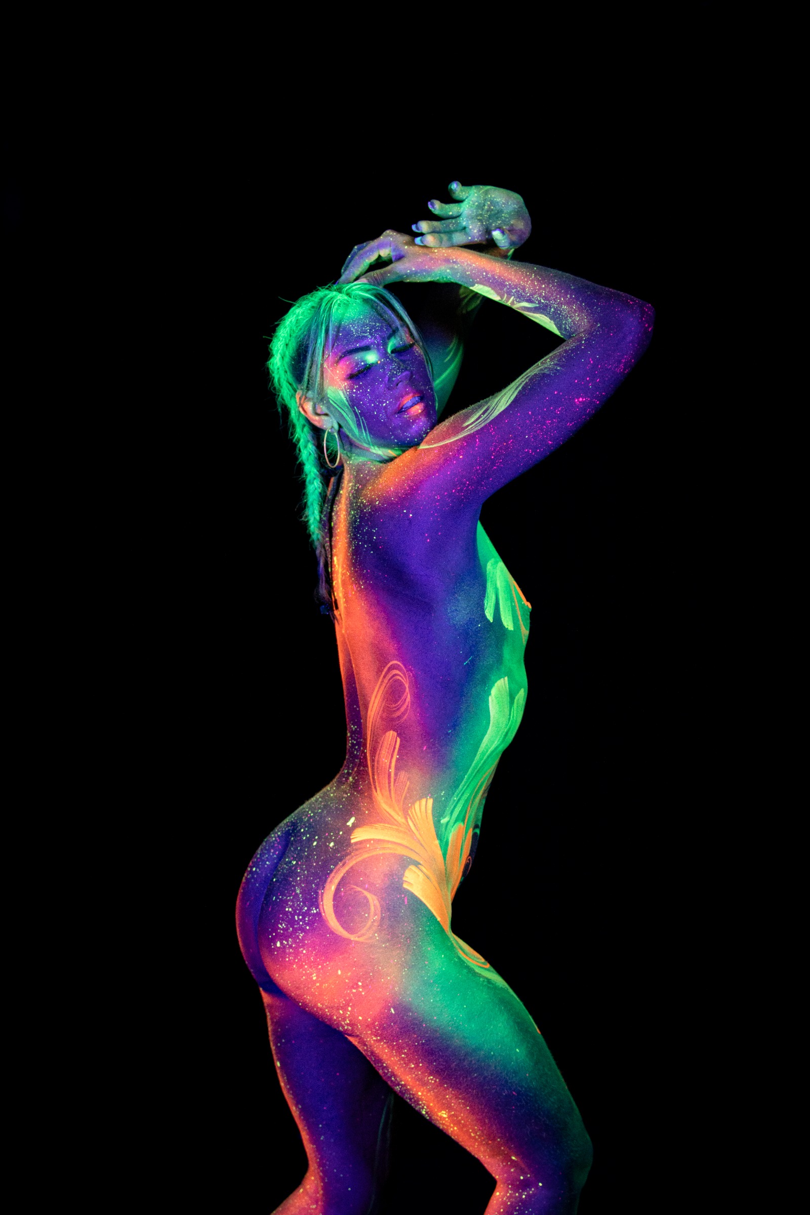 Miss_Layla_X stands facing three-quarters away from the camera, her body painted in purple, orange, and green UV tones with a swirl design on her lower back. Bodypaint by Claudia Kraemer. Photographed by Elad Itzkin Photography under UV blacklight in his studio in Gesundbrunnen, Berlin.
