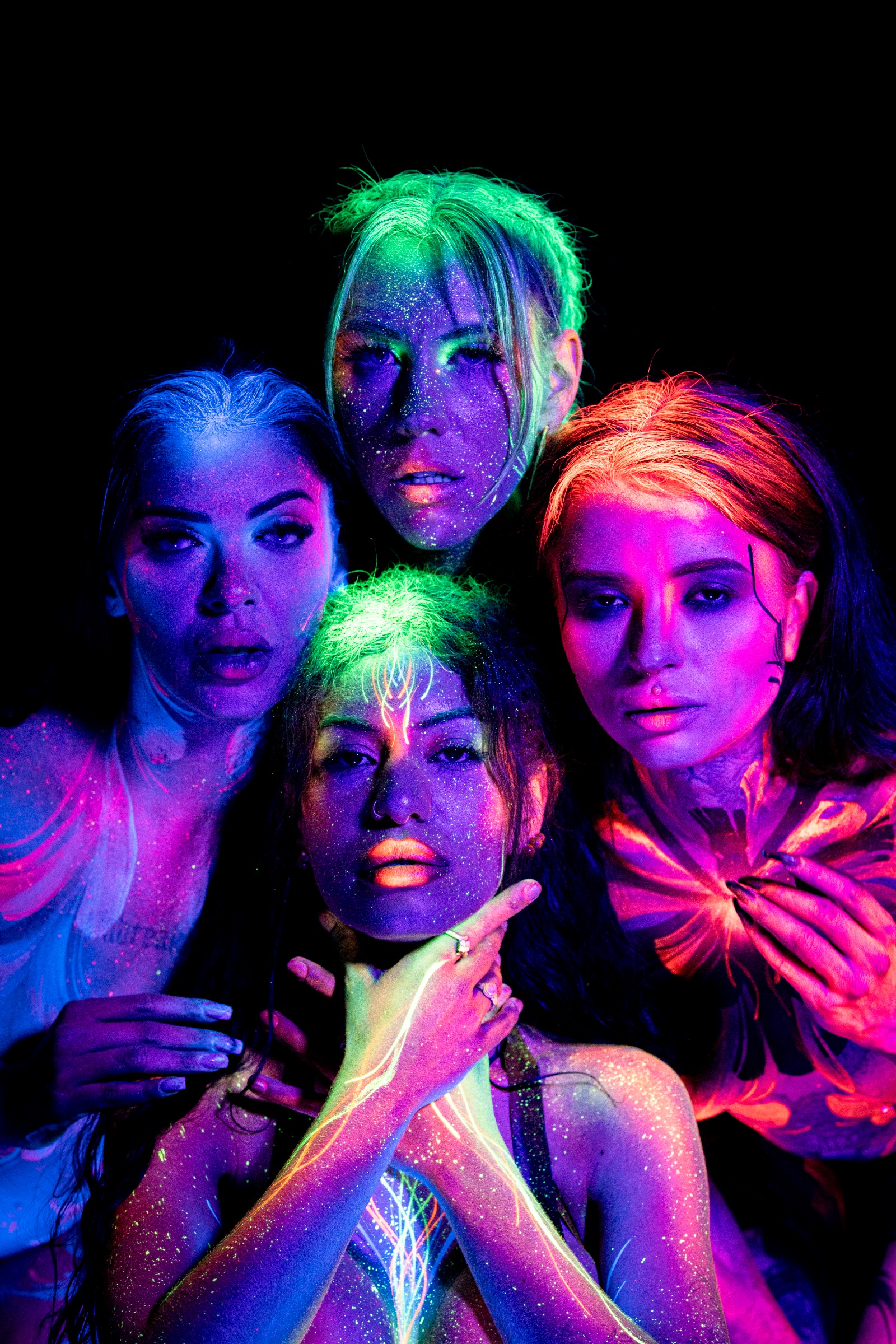 Models PinkMephisto, AlyssaHotofficial, BabeyGianna, LadySnke, and Miss_Layla_X are photographed close together, several standing with one kneeling in front, faces gathered close to the camera. Their UV bodypaint and makeup appear in blue, green, and orange tones. Bodypaint by Claudia Kraemer. Photographed by Elad Itzkin Photography under UV blacklight in his studio in Gesundbrunnen, Berlin.