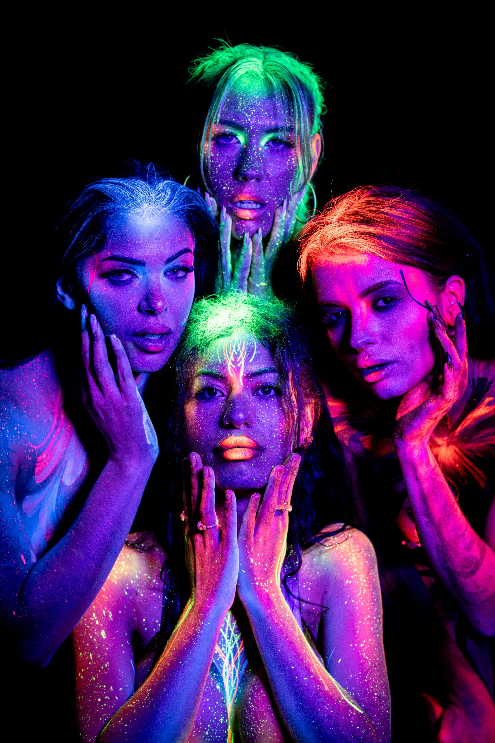 PinkMephisto, AlyssaHotofficial, BabeyGianna, LadySnke, and Miss_Layla_X pose closely together, most standing with one kneeling in the front of the group, faces close to the lens. UV bodypaint and makeup span blue, green, and orange tones. Bodypaint by Claudia Kraemer. Photographed by Elad Itzkin Photography under UV blacklight in his studio in Gesundbrunnen, Berlin.