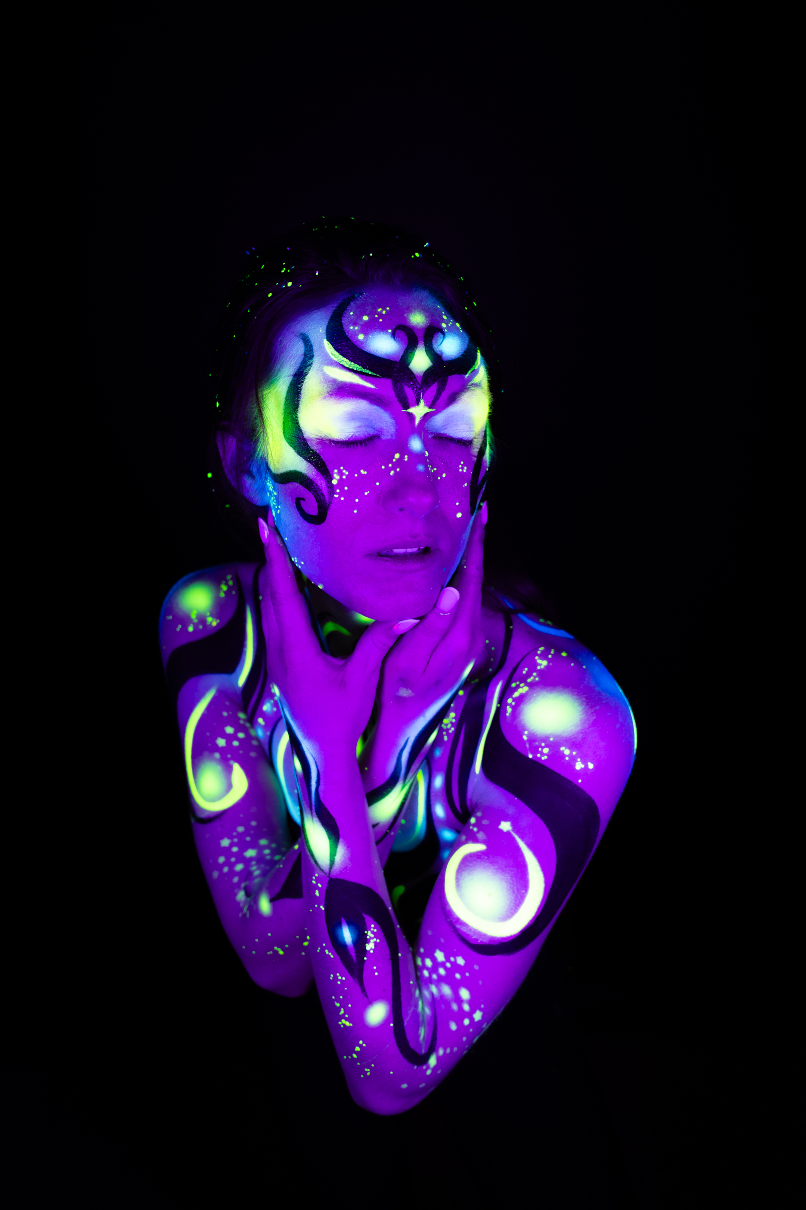 Sage is framed from the chest up, head tilted back in a three-quarter view. The bodypaint is a geometric design in purple, blue, and green tones, with a crown-like pattern across the forehead. Bodypaint by Joana Rohr. Photographed by Elad Itzkin Photography under UV light in his studio in Gesundbrunnen, Berlin.