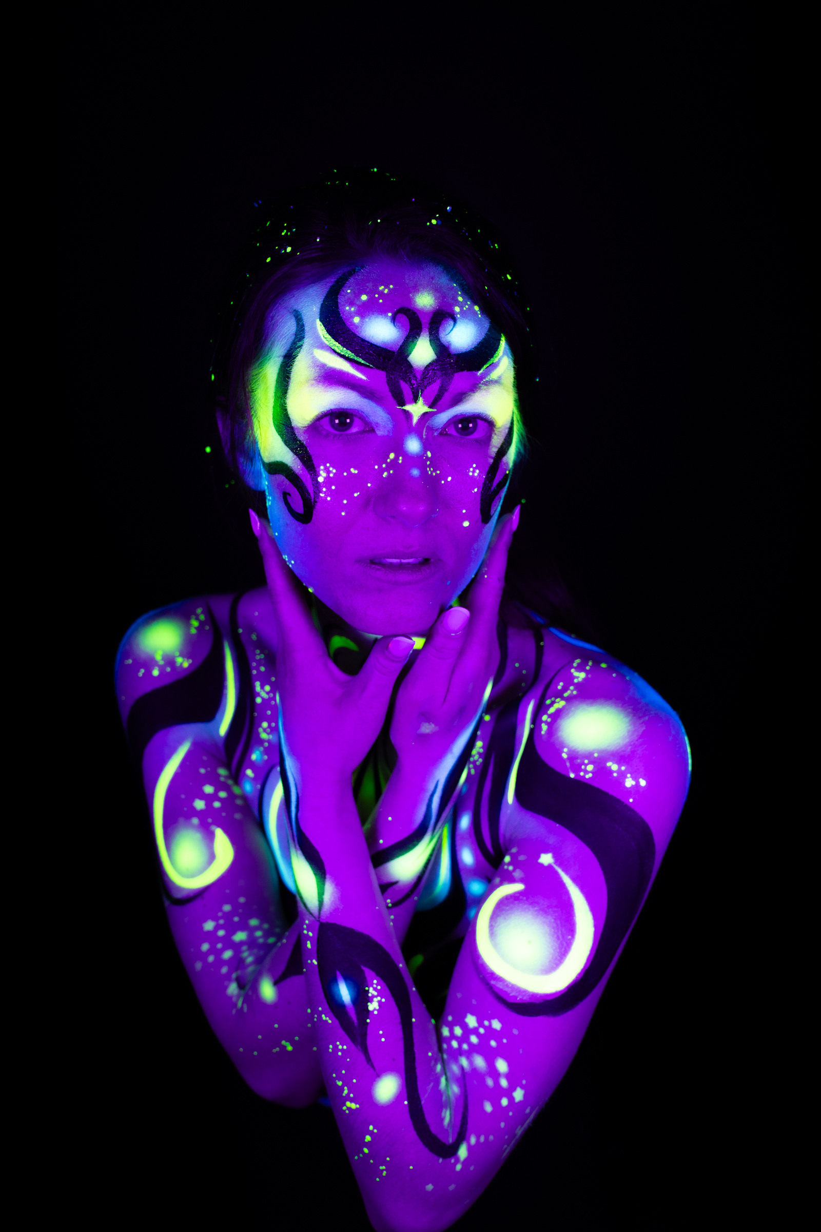 Sage is framed from the chest up, facing forward directly. The bodypaint design combines purple, blue, and green geometric linework across the face, neck, and shoulders. Bodypaint by Joana Rohr. Photographed by Elad Itzkin Photography under UV light in his studio in Gesundbrunnen, Berlin.