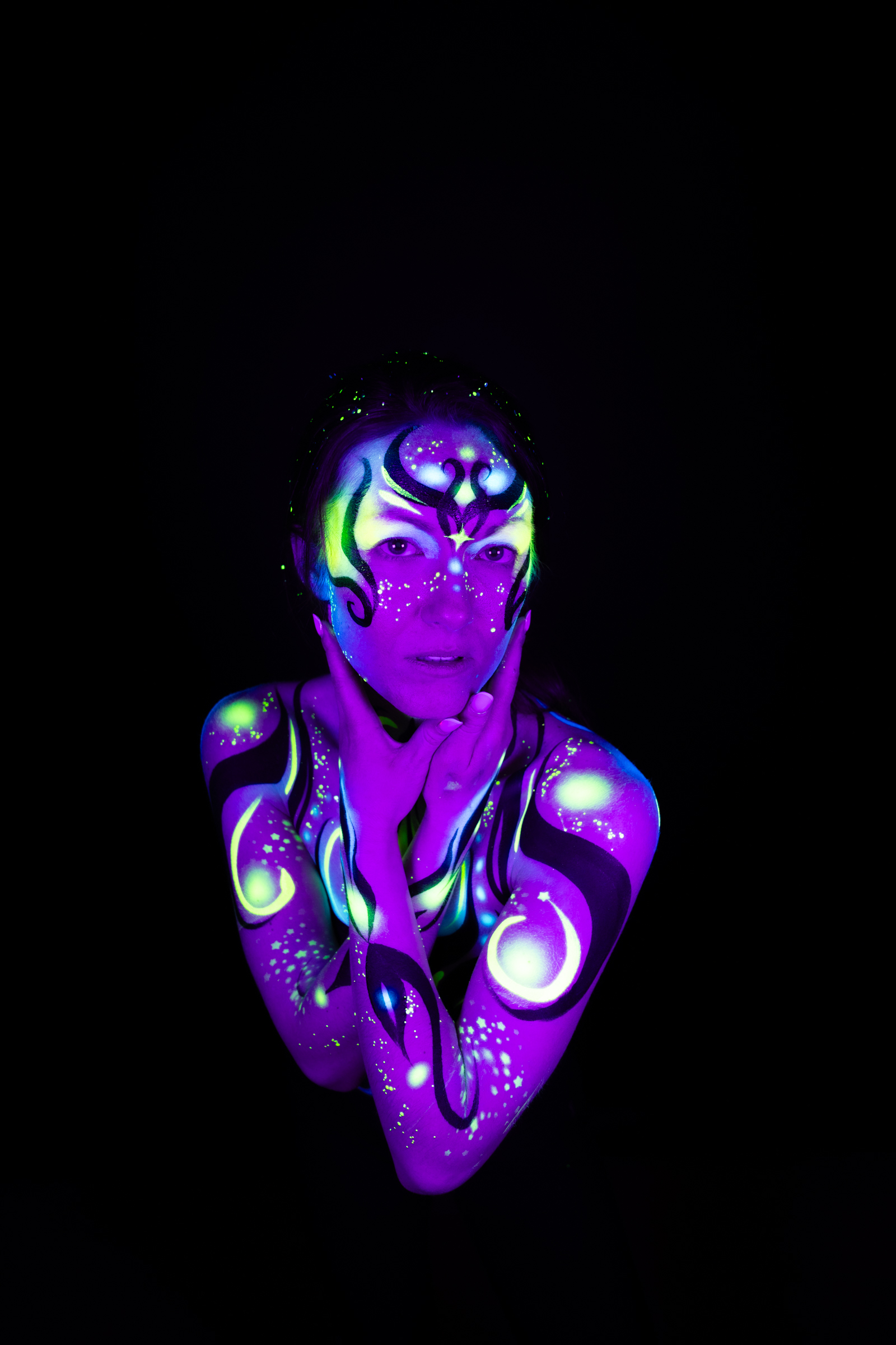 Sage stands facing the camera, shown from the chest up in a slightly wider frame. Her face and upper body are painted with a purple and blue geometric pattern accented in green, with a symmetrical motif centered on the forehead. Bodypaint by Joana Rohr. Photographed by Elad Itzkin Photography under UV light in his studio in Gesundbrunnen, Berlin.
