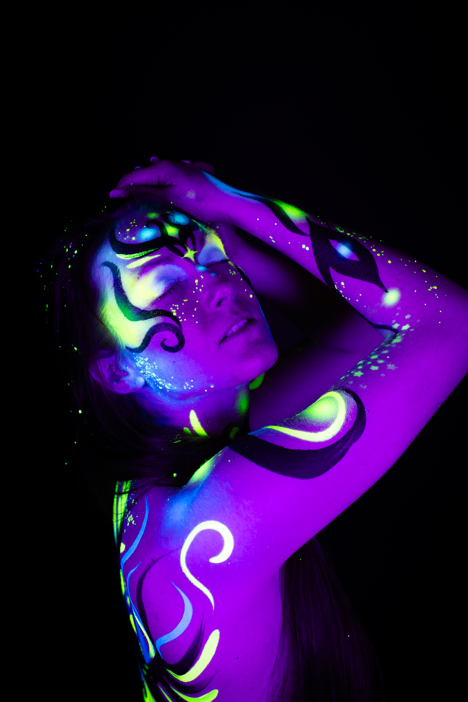 Sage stands with her head tipped back, face turned upward and to the side. Purple and blue abstract linework with green accents covers her face, neck, and upper torso. Bodypaint by Joana Rohr. Photographed by Elad Itzkin Photography under UV/blacklight in his studio in Gesundbrunnen, Berlin.