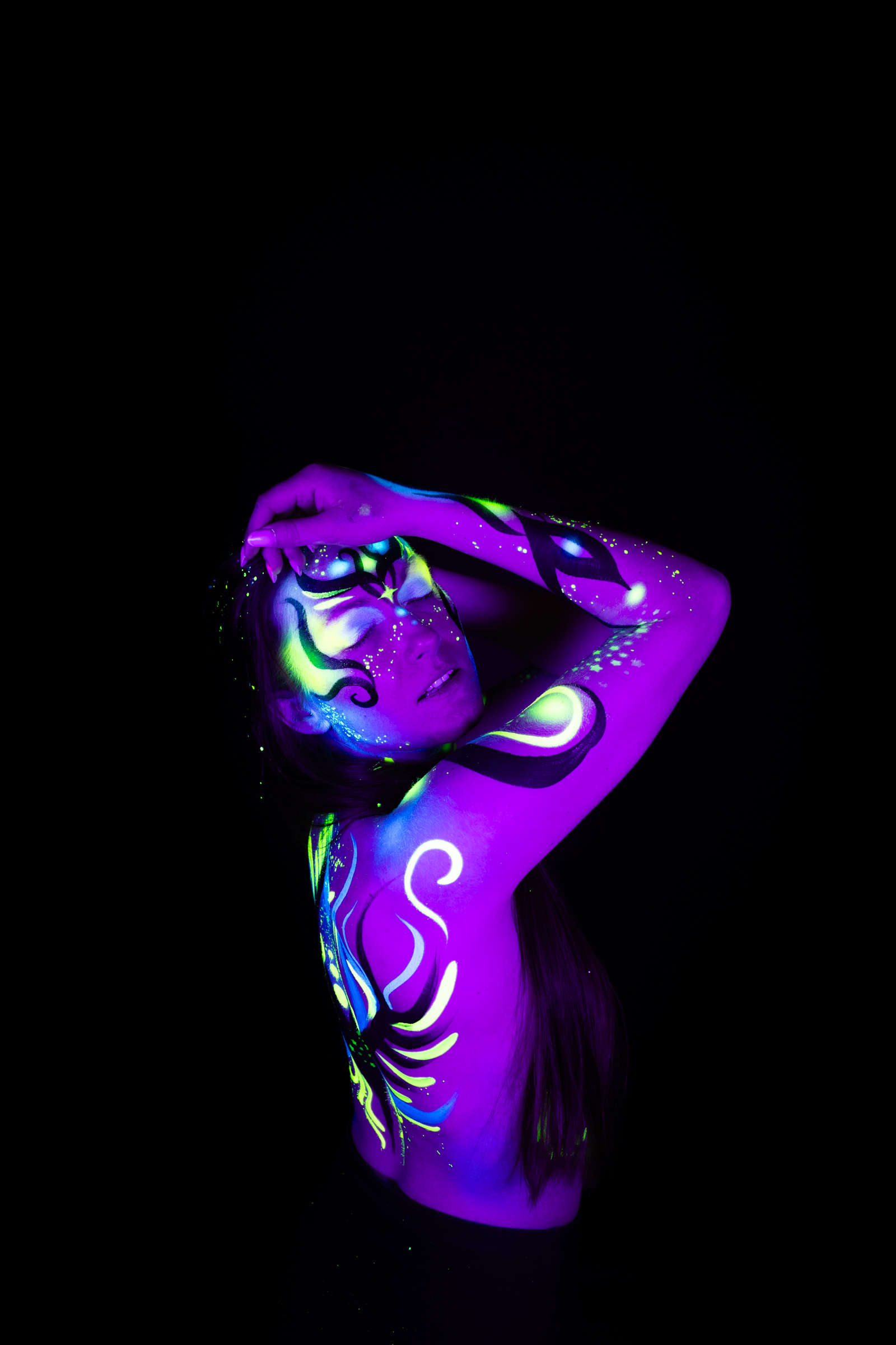 Sage stands with her head tilted back in a three-quarter pose. A swirling abstract design in purple, blue, and green spans her face, neck, and upper chest. Bodypaint by Joana Rohr. Photographed by Elad Itzkin Photography under UV/blacklight in his studio in Gesundbrunnen, Berlin.