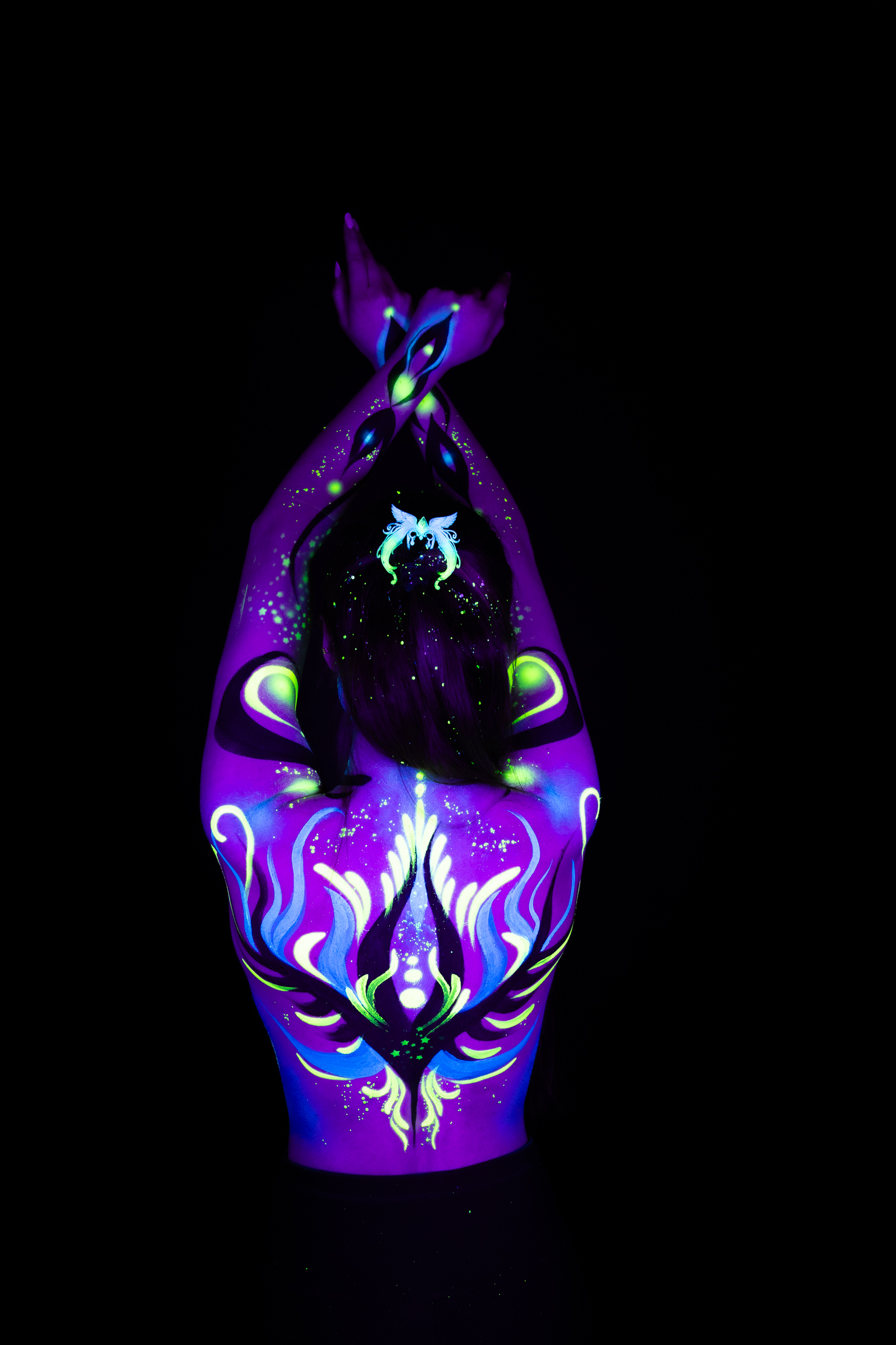 Sage stands facing the camera in a tight torso crop. Layered purple and blue swirling shapes with green accents form a silhouette pattern centered on the chest. Bodypaint by Joana Rohr. Photographed by Elad Itzkin Photography under UV/blacklight in his studio in Gesundbrunnen, Berlin.
