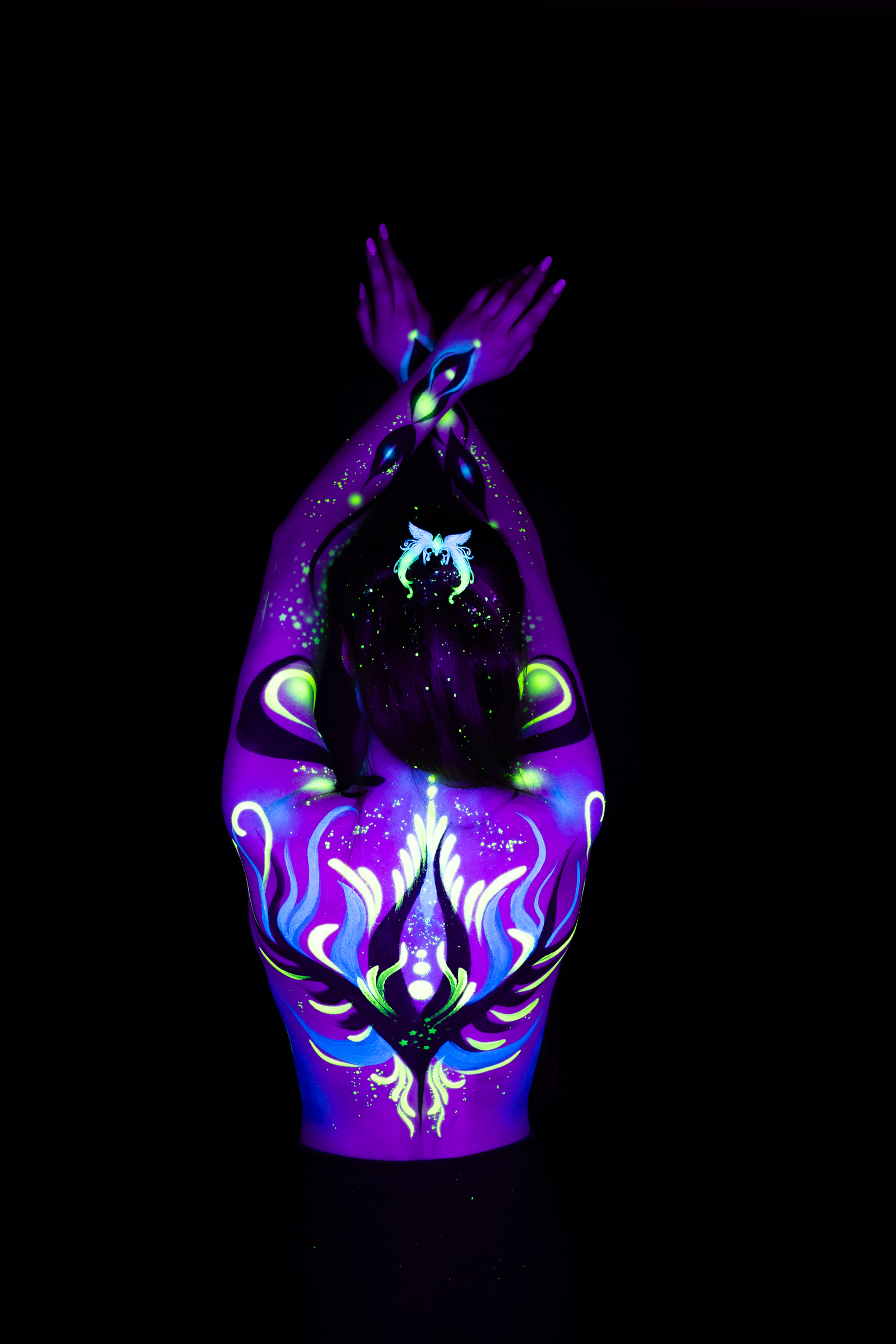 Sage stands with her back to the camera, her head lowered slightly. A symmetrical design in purple, blue, and green spans her upper back and shoulders. Bodypaint by Joana Rohr. Photographed by Elad Itzkin Photography under UV/blacklight in his studio in Gesundbrunnen, Berlin.