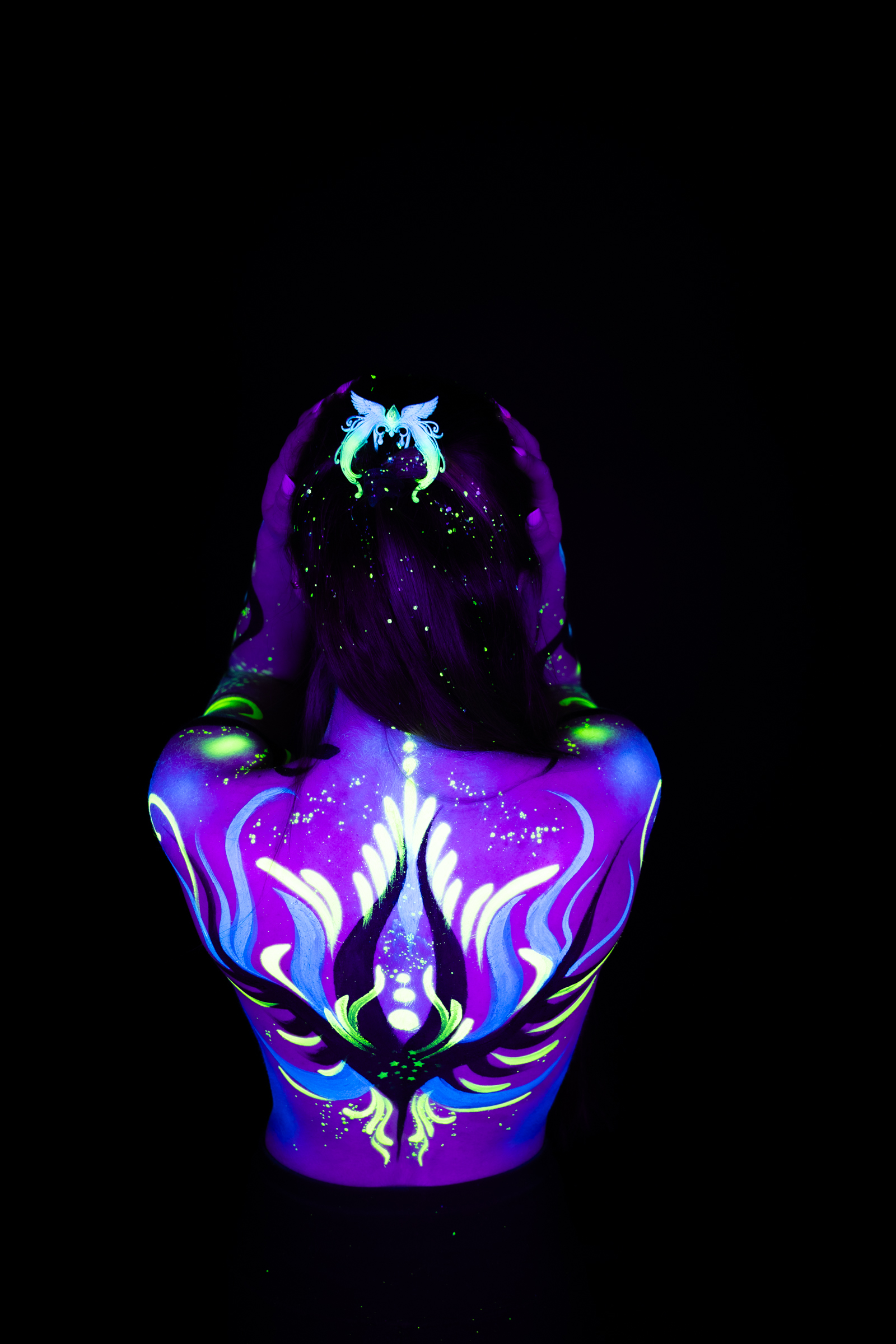 Sage stands with her back turned to the camera in a close upper-body crop. Purple, blue, and green abstract linework forms a symmetrical pattern across her shoulders and upper back. Bodypaint by Joana Rohr. Photographed by Elad Itzkin Photography under UV/blacklight in his studio in Gesundbrunnen, Berlin.