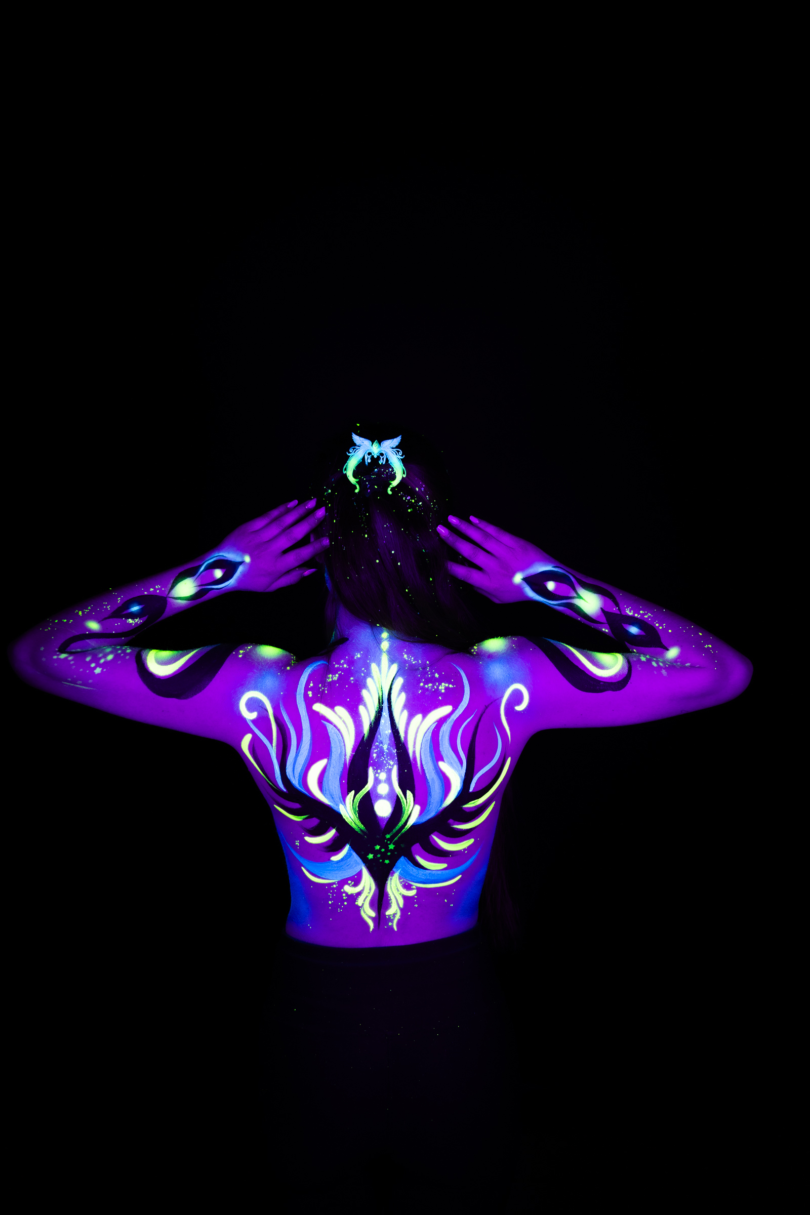 Standing with her back turned, Sage is shown in a close upper-body crop. Purple and blue tones form a symmetrical design across her shoulders and upper back, accented with green. Bodypaint by Joana Rohr. Photographed by Elad Itzkin Photography under UV/blacklight in his studio in Gesundbrunnen, Berlin.
