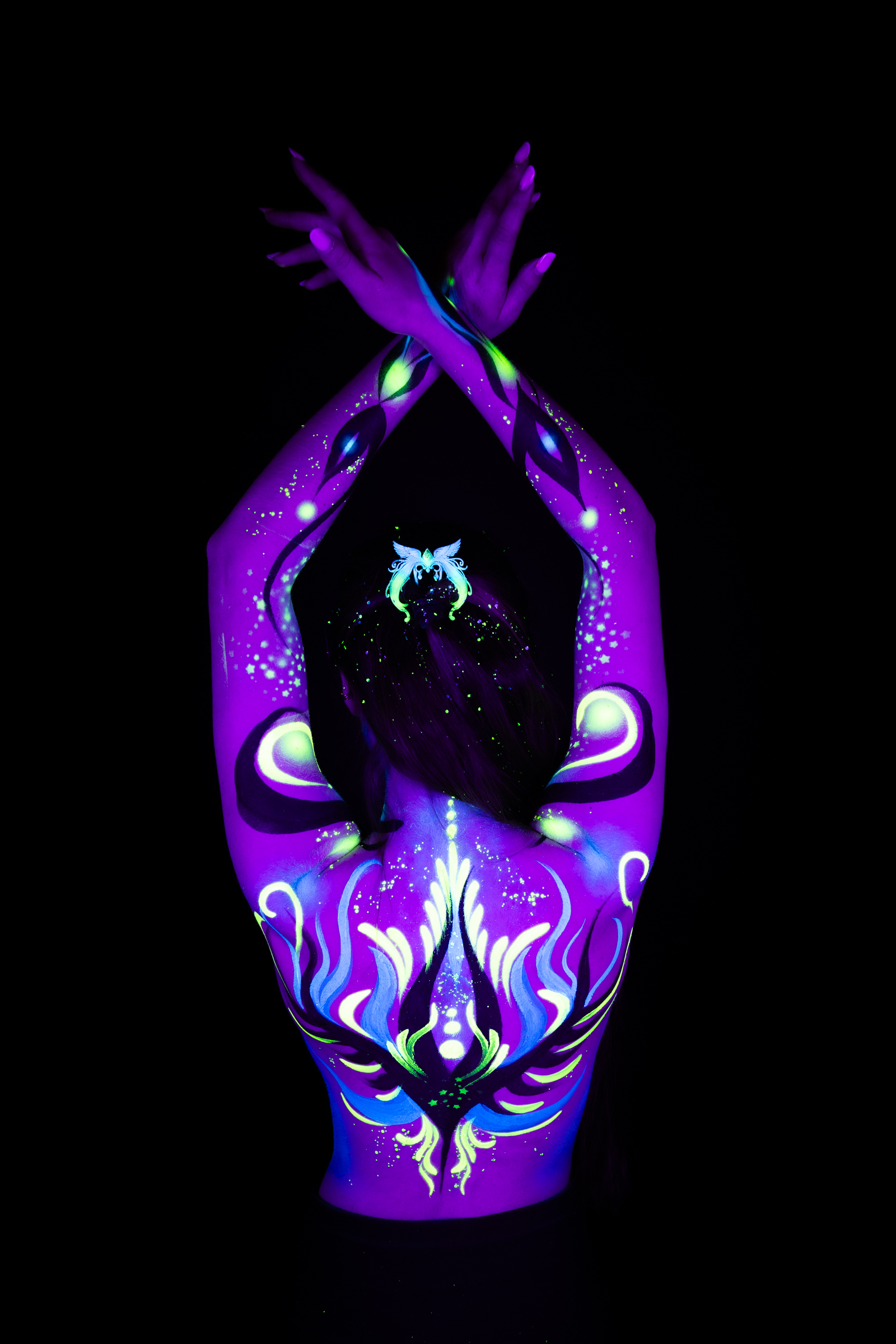 Sage stands with her back to the camera in a tight crop of the upper body. A symmetrical abstract design in purple, blue, and green covers her shoulders and back. Bodypaint by Joana Rohr. Photographed by Elad Itzkin Photography under UV/blacklight in his studio in Gesundbrunnen, Berlin.