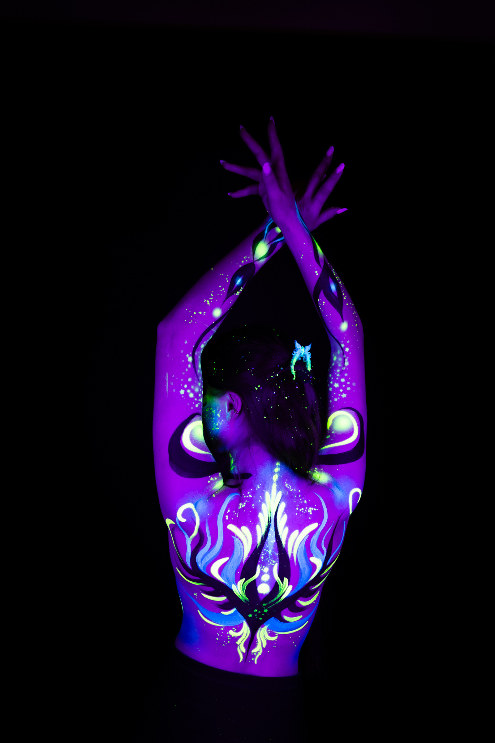 Sage stands with her back to the camera and her head bowed slightly forward. A symmetrical floral pattern in purple, blue, and green spans her shoulders and upper back. Bodypaint by Joana Rohr. Photographed by Elad Itzkin Photography under UV/blacklight in his studio in Gesundbrunnen, Berlin.