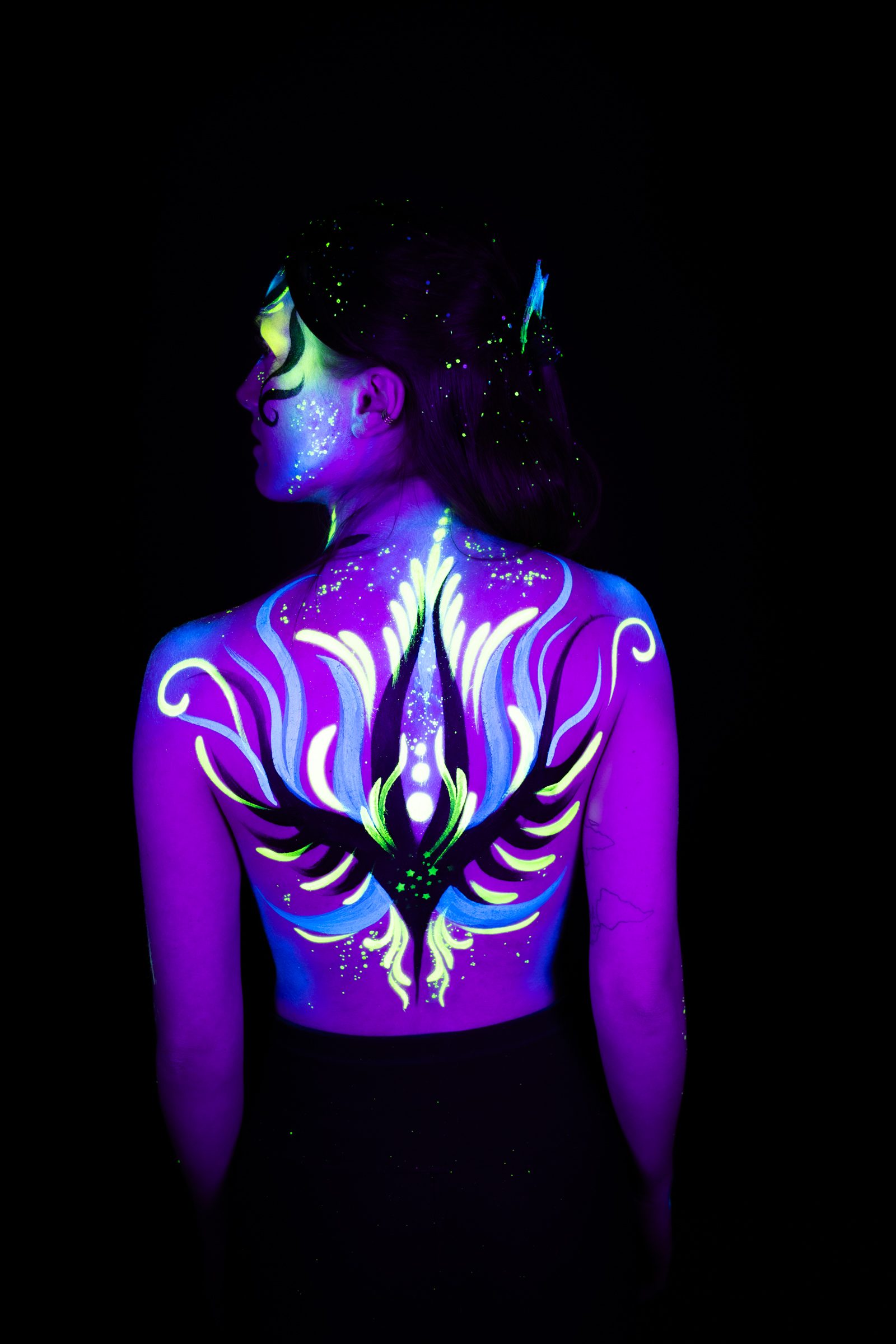 Sage stands with her back to the camera, her face turned to the side in profile. Purple, blue, and green abstract linework forms a symmetrical pattern across her upper back. Bodypaint by Joana Rohr. Photographed by Elad Itzkin Photography under UV/blacklight in his studio in Gesundbrunnen, Berlin.