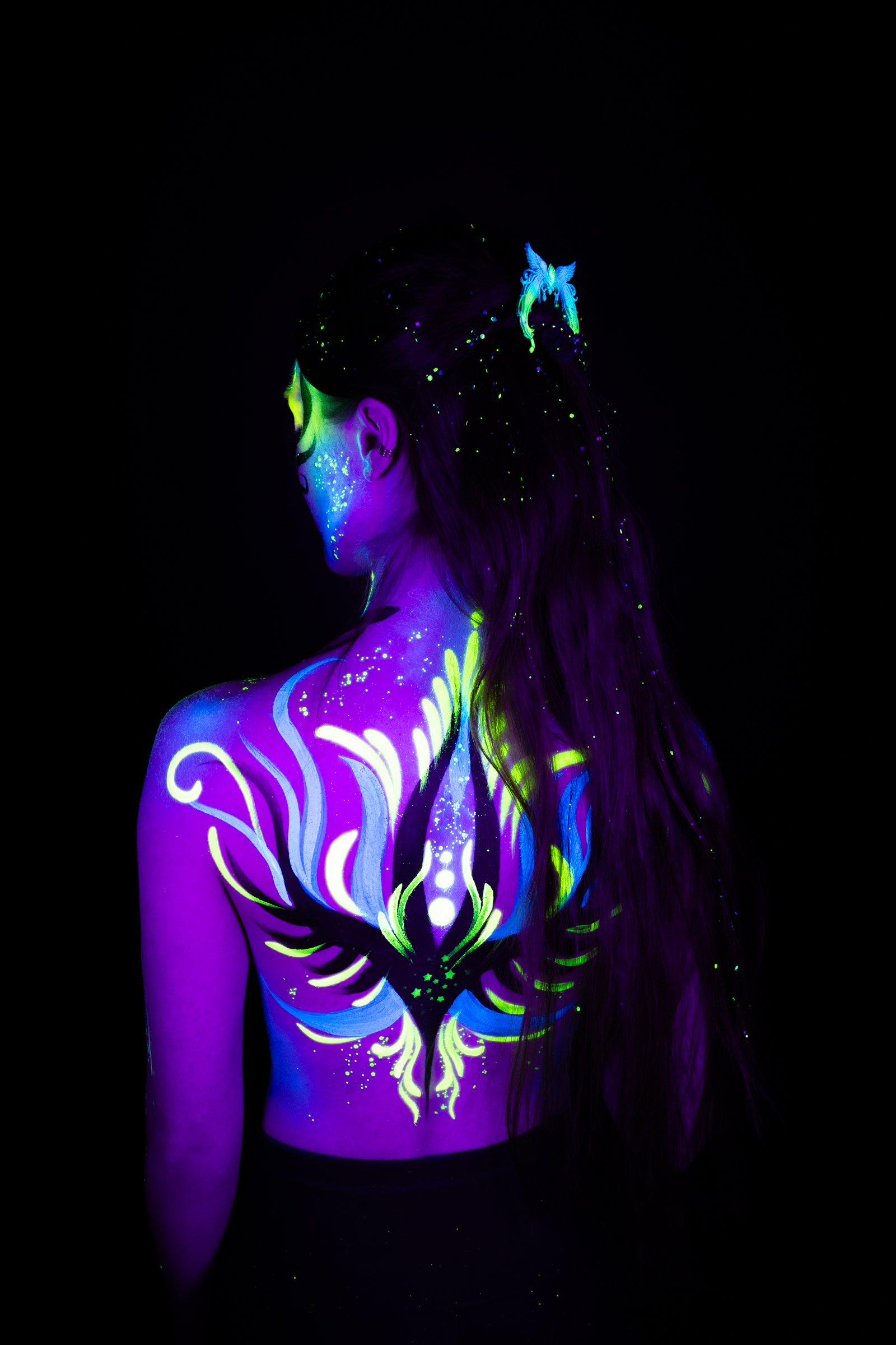 Sage stands with her back to the camera and her head turned to one side in profile. The bodypaint forms a symmetrical purple, blue, and green pattern across her upper back. Bodypaint by Joana Rohr. Photographed by Elad Itzkin Photography under UV/blacklight in his studio in Gesundbrunnen, Berlin.