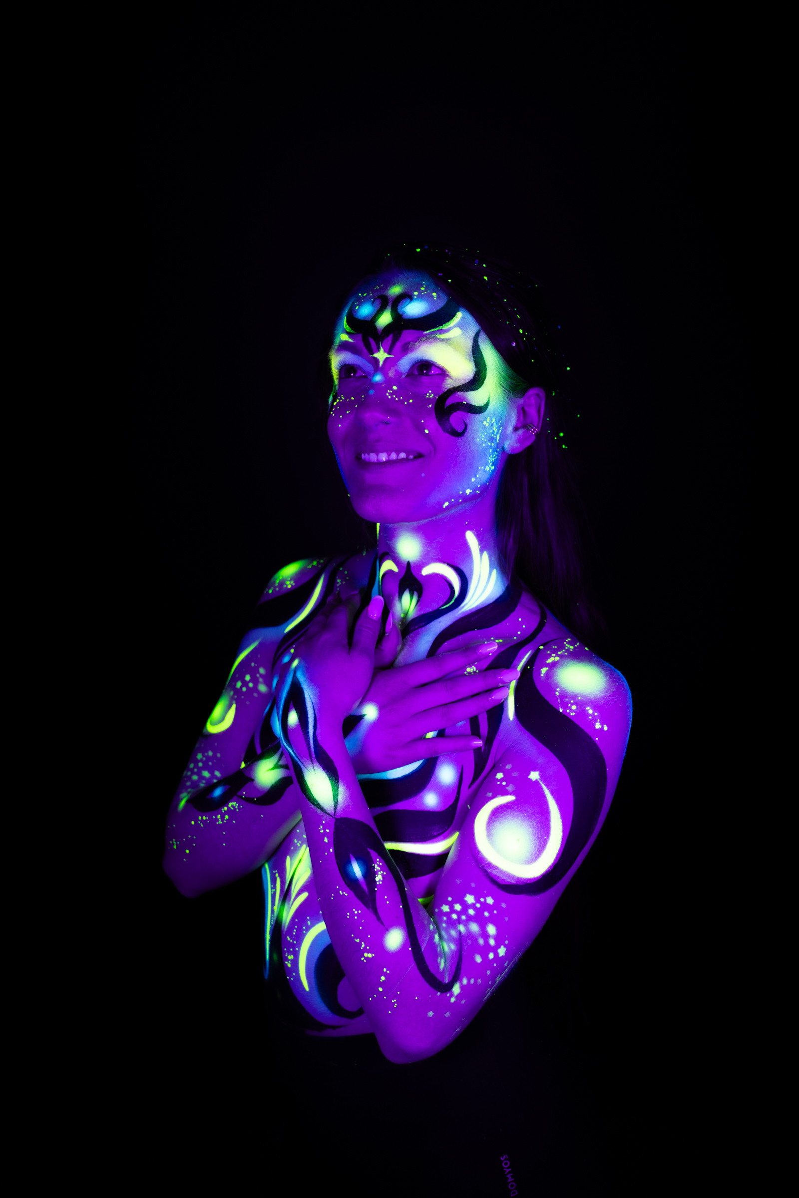 Facing the camera in a three-quarter pose, Sage displays a swirling design in purple, blue, and green abstract linework covering her face and chest. Bodypaint by Joana Rohr. Photographed by Elad Itzkin Photography under UV/blacklight in his studio in Gesundbrunnen, Berlin.