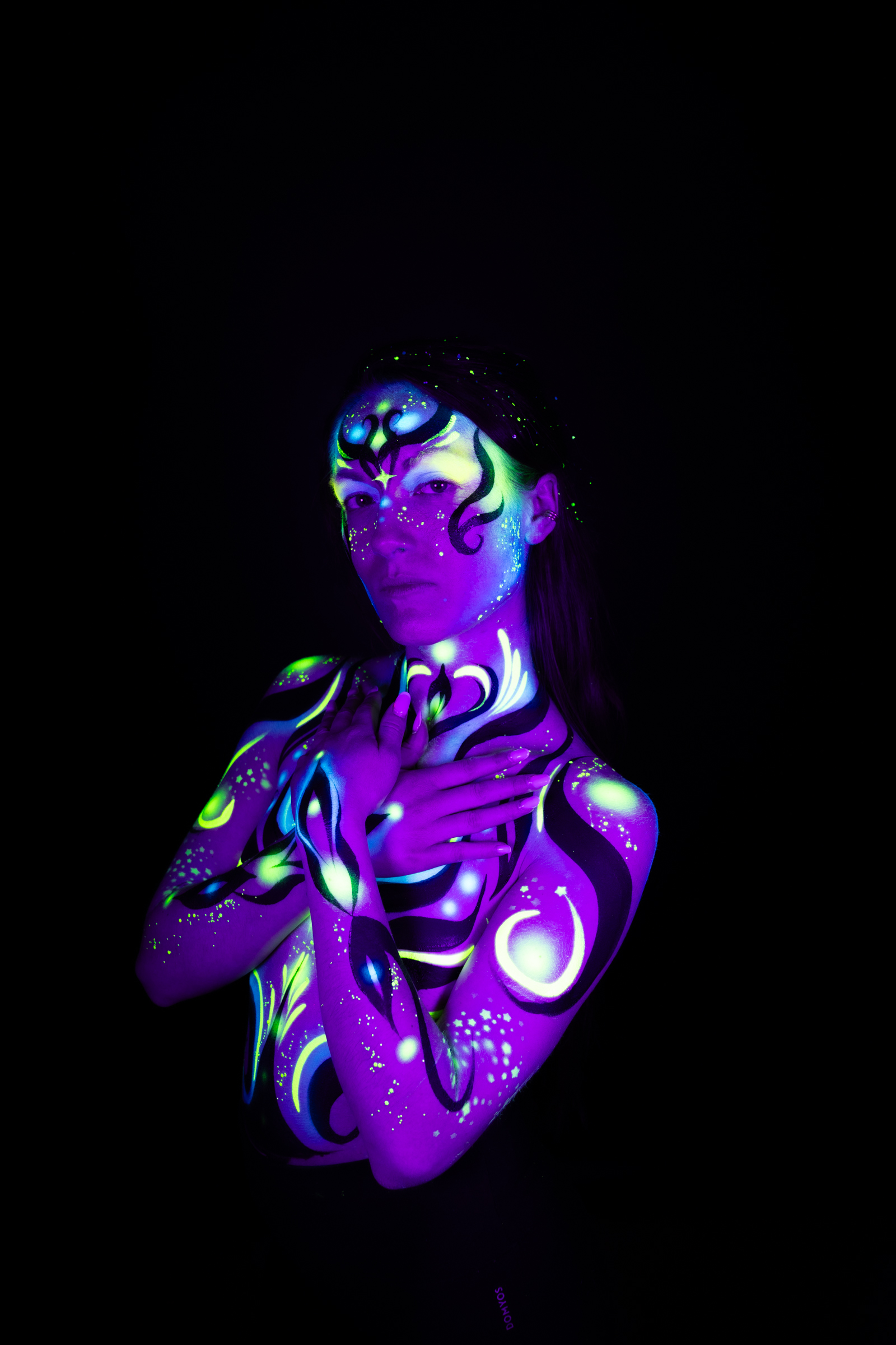 Sage stands in a three-quarter pose with her face turned to the side. The design combines purple, blue, and green in an abstract swirling pattern across her face and chest. Bodypaint by Joana Rohr. Photographed by Elad Itzkin Photography under UV/blacklight in his studio in Gesundbrunnen, Berlin.