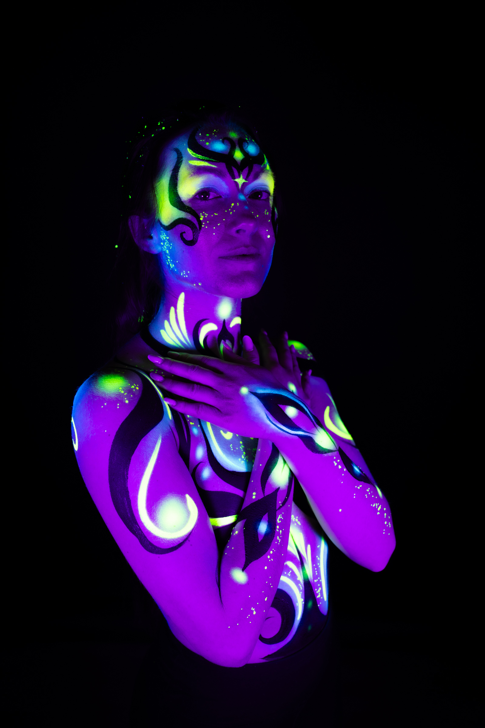 Sage stands in a three-quarter view with her head turned to one side, her face and chest marked with a purple, blue, and green UV-reactive floral and geometric design. Bodypaint by Joana Rohr, photographed by Elad Itzkin Photography under UV light in his studio in Gesundbrunnen, Berlin.