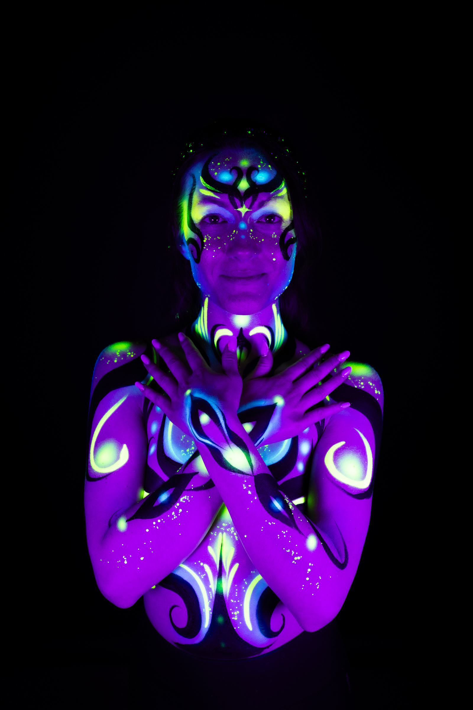 Sage faces the camera directly in a frontal standing pose, her chest and shoulders covered in a symmetrical purple, blue, and green UV-reactive floral pattern radiating outward from the center. Bodypaint by Joana Rohr, photographed by Elad Itzkin Photography under UV light in his studio in Gesundbrunnen, Berlin.