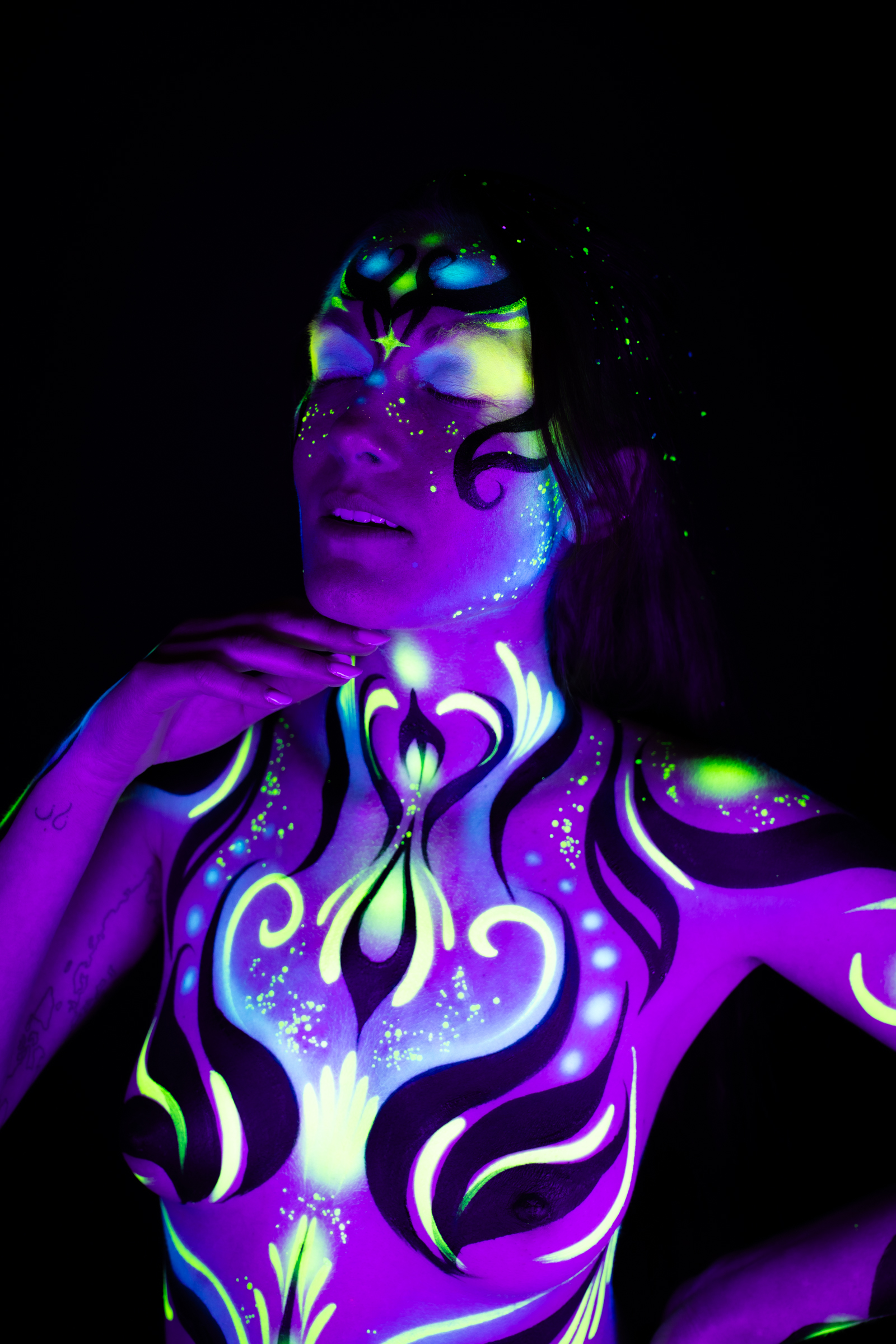 Sage stands with her head tilted back in a three-quarter view, her torso decorated with a purple, blue, and green UV-reactive floral design featuring swirling linework and star-shaped accents. Bodypaint by Joana Rohr, photographed by Elad Itzkin Photography under UV light in his studio in Gesundbrunnen, Berlin.