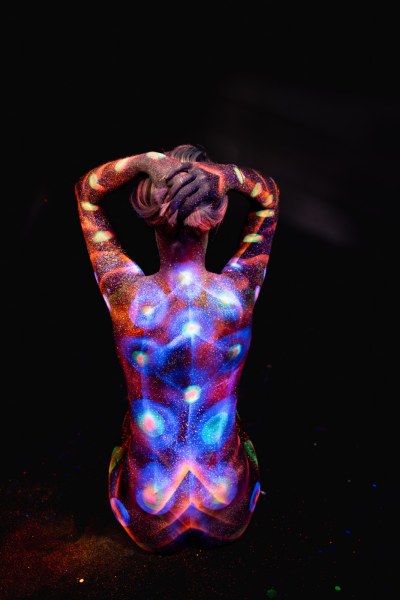 Selina kneels with her back to the camera. Her body is covered in a symmetrical UV bodypaint design combining orange, purple and blue tones with a glittering, dotted texture. Bodypaint by Elad Itzkin. Photographed by Elad Itzkin Photography under UV light in his studio in Gesundbrunnen, Berlin.