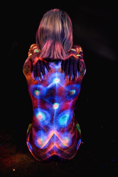 Kneeling with her back turned to the camera, Selina is painted in a symmetrical mandala-like UV design in orange, blue and purple with a sparkling, dotted texture. Bodypaint by Elad Itzkin. Photographed by Elad Itzkin Photography under blacklight in his studio in Gesundbrunnen, Berlin.