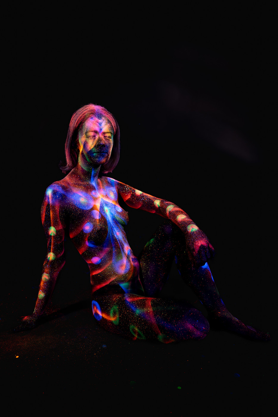 Selina sits in a three-quarter pose, her body painted with a UV design of orange, blue and green stripes and dots that give a glittering, textured appearance. Bodypaint by Elad Itzkin. Photographed by Elad Itzkin Photography under UV light in his studio in Gesundbrunnen, Berlin.