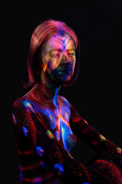 Seated in a three-quarter pose, Selina's torso and legs are marked with a colorful UV bodypaint pattern of stripes, dots and starburst-like linework in orange, blue and green. Bodypaint by Elad Itzkin. Photographed by Elad Itzkin Photography under blacklight in his studio in Gesundbrunnen, Berlin.
