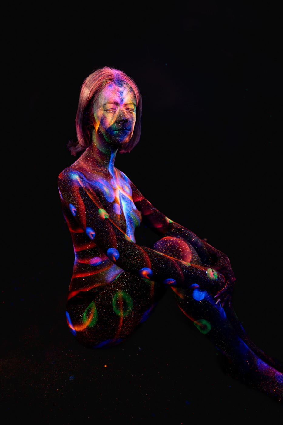 Seated with one leg drawn up, Selina's torso is painted with a UV design of orange and blue linework, with scattered dot patterns across the shoulders and chest. Bodypaint by Elad Itzkin. Photographed by Elad Itzkin Photography under UV light in his studio in Gesundbrunnen, Berlin.