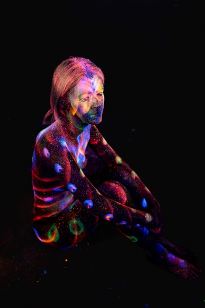 Selina sits in profile with one knee raised, her torso and legs covered in a UV bodypaint pattern of orange stripes and scattered dots. Bodypaint by Elad Itzkin. Photographed by Elad Itzkin Photography under blacklight in his studio in Gesundbrunnen, Berlin.